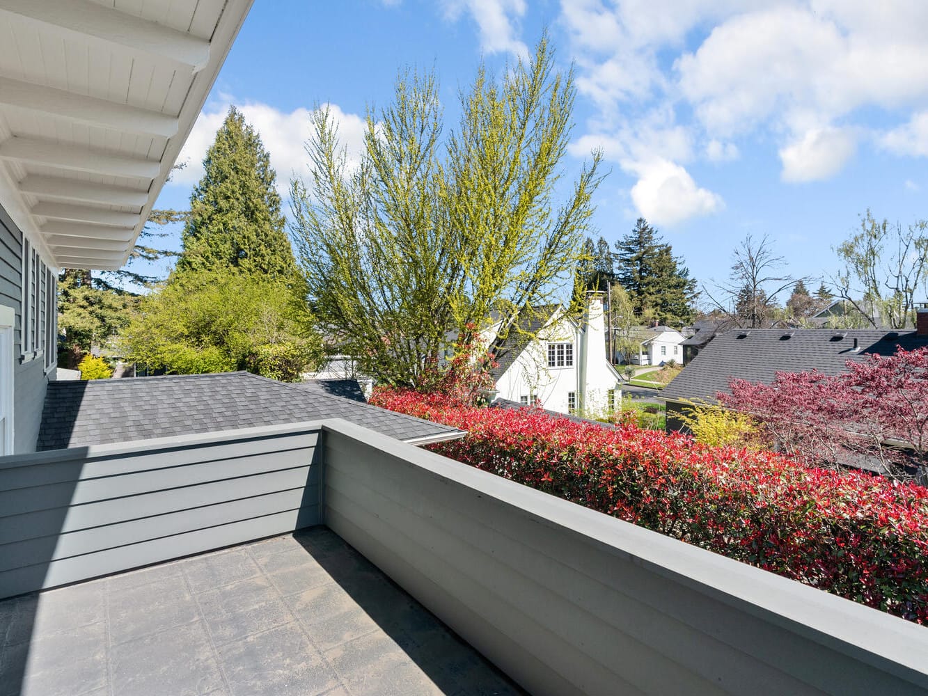 View from a gray house balcony overlooking a suburban neighborhood. The scene includes a roof, trees, and houses under a blue sky with scattered clouds. Red and green foliage lines the edge of the balcony.