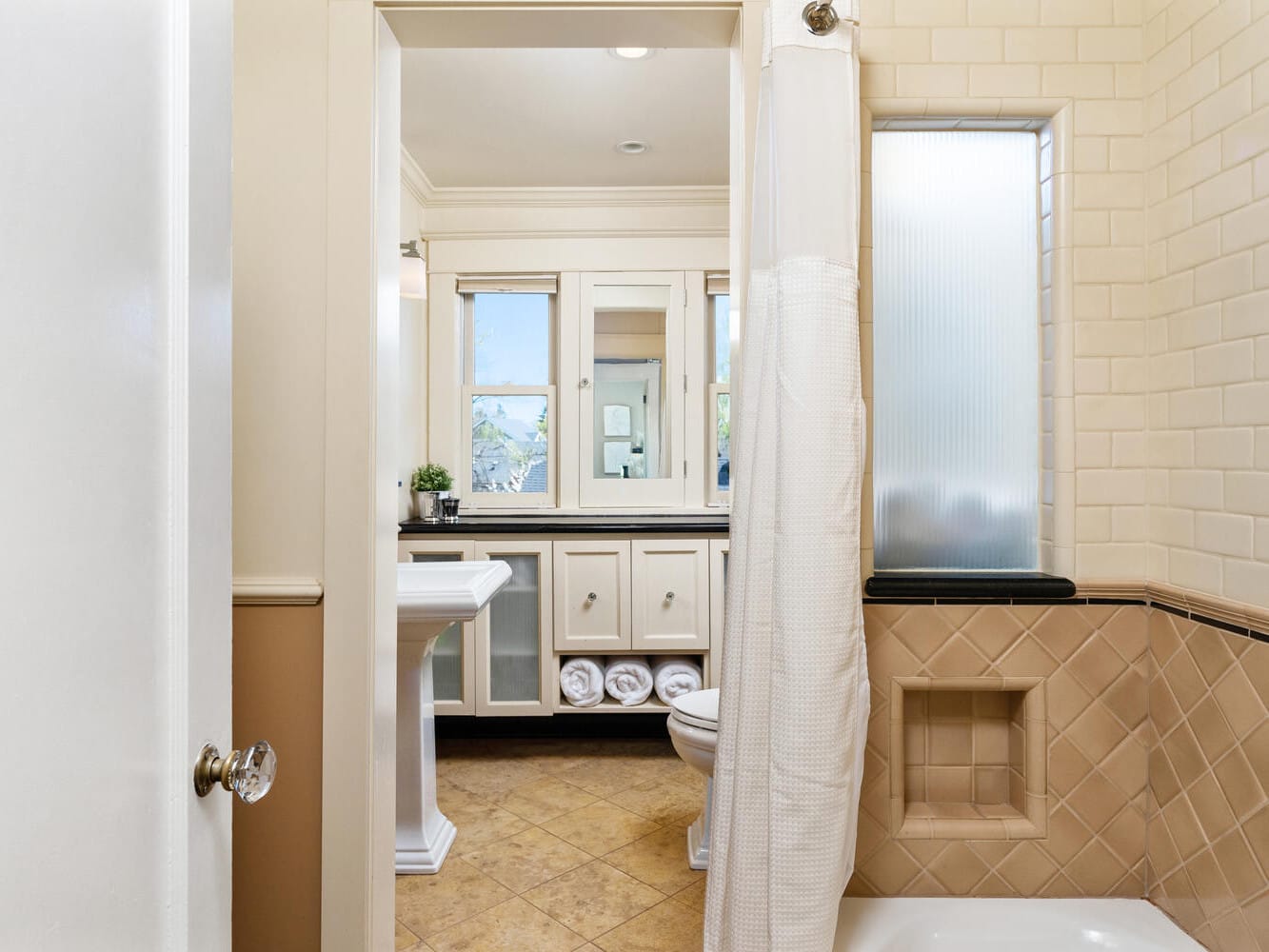 A bathroom with beige tiled walls, a white pedestal sink, and a shower with a curtain. The window is frosted, and theres a narrow shelf in the shower area. The floor has brown tiles, and theres storage space beneath the sink with rolled towels.