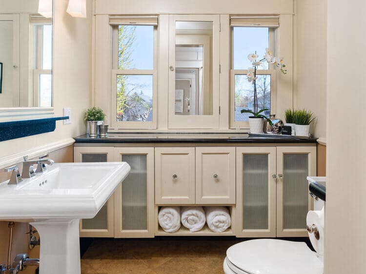 A bright bathroom with a pedestal sink and a toilet on a tiled floor. Cabinets under the window hold rolled towels. A mirror reflects natural light from the window, and there are small plants on the counter.