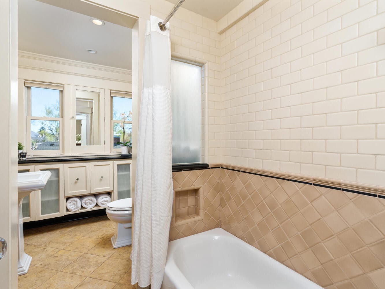 A bathroom with a white bathtub, beige tiled walls, and cream floor tiles. A white shower curtain is partly open. The vanity area features a sink, toilet, and wall mirror with cabinets beneath. Natural light comes in through two windows.