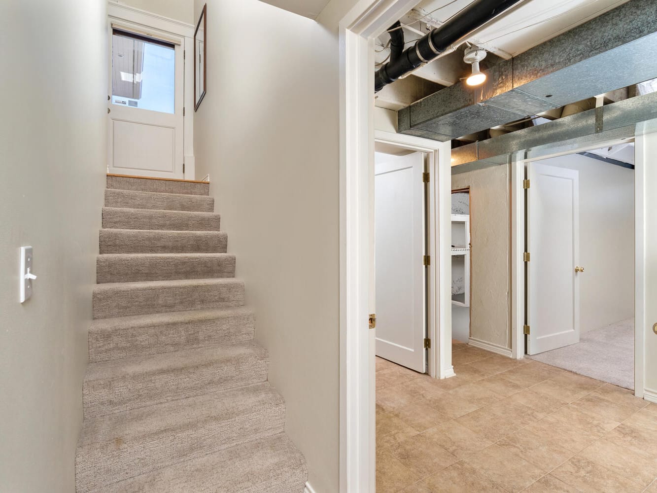 A basement hallway with carpeted stairs leading up to a door with a window. Two open doorways on the right reveal rooms with light-colored walls and tiled flooring. Exposed ceiling pipes are visible.