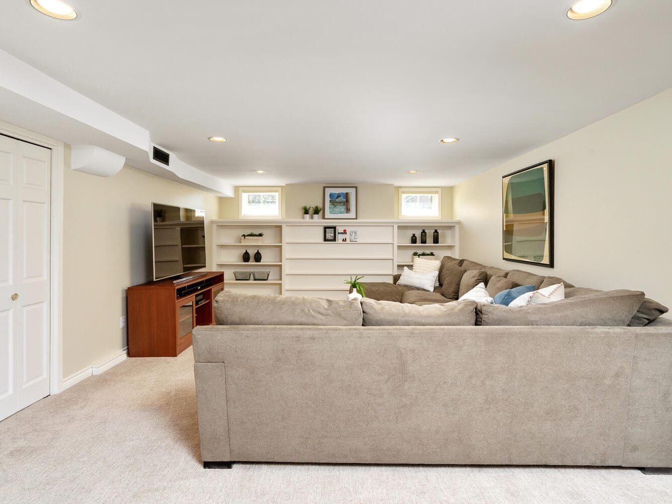 A basement living room with beige walls and carpet, featuring a large L-shaped brown sectional sofa. Theres a wooden TV stand with a flat-screen TV, shelves with decor items, and framed art on the walls. Small windows let in natural light.