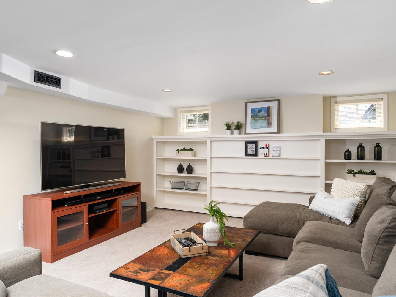 A cozy living room with a large sectional sofa and a colorful coffee table. A flat-screen TV sits on a wooden stand, and built-in white shelves with decor items are on the wall behind. Two small windows bring in natural light.