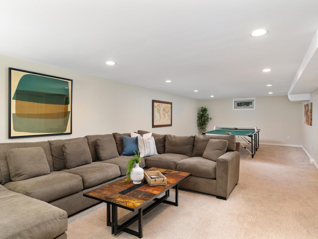 Spacious basement room with beige walls and carpet, featuring a large brown sectional sofa and a rectangular coffee table. A ping pong table is visible in the background. Wall art includes abstract and world map images. Ceiling lights illuminate the space.