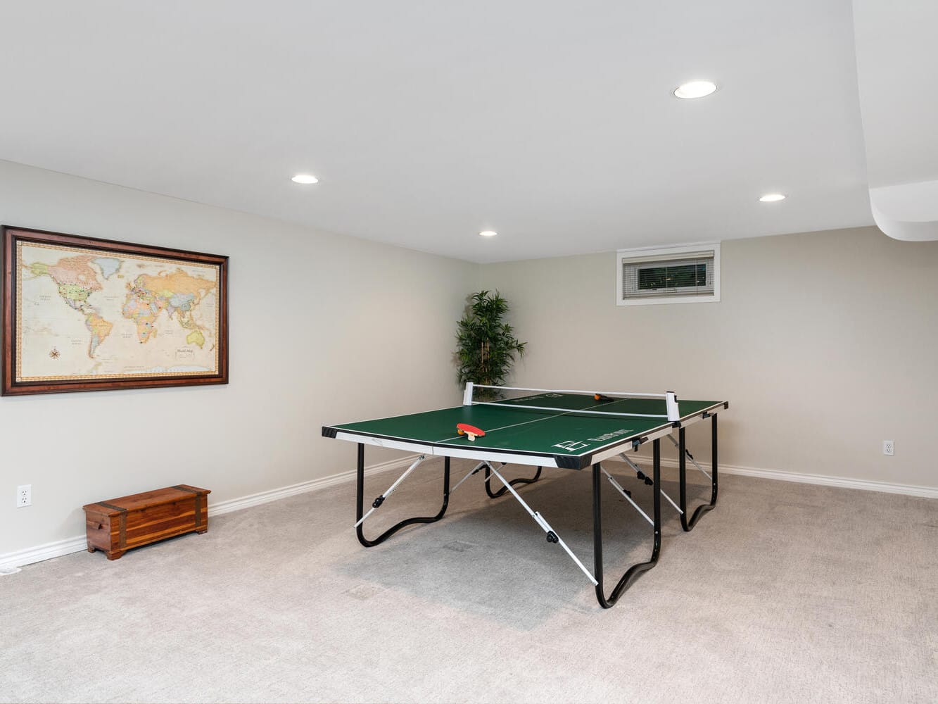 A basement room with beige walls features a ping pong table at the center. A world map is hung on the wall to the left, and a small window is near the ceiling. A plant adds greenery in the corner, and a wooden chest sits under the map.