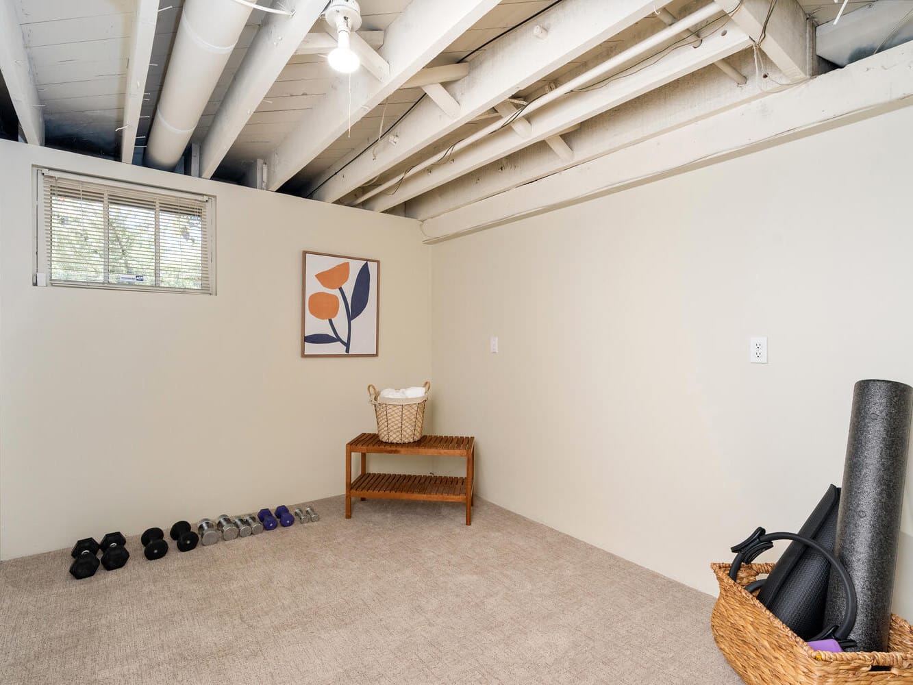 A small exercise room with dumbbells and weights lined up on the carpet. A wicker basket on a wooden shelf holds a towel. Yoga mats are rolled up in a wicker basket nearby. A window and abstract art adorn the pale walls.