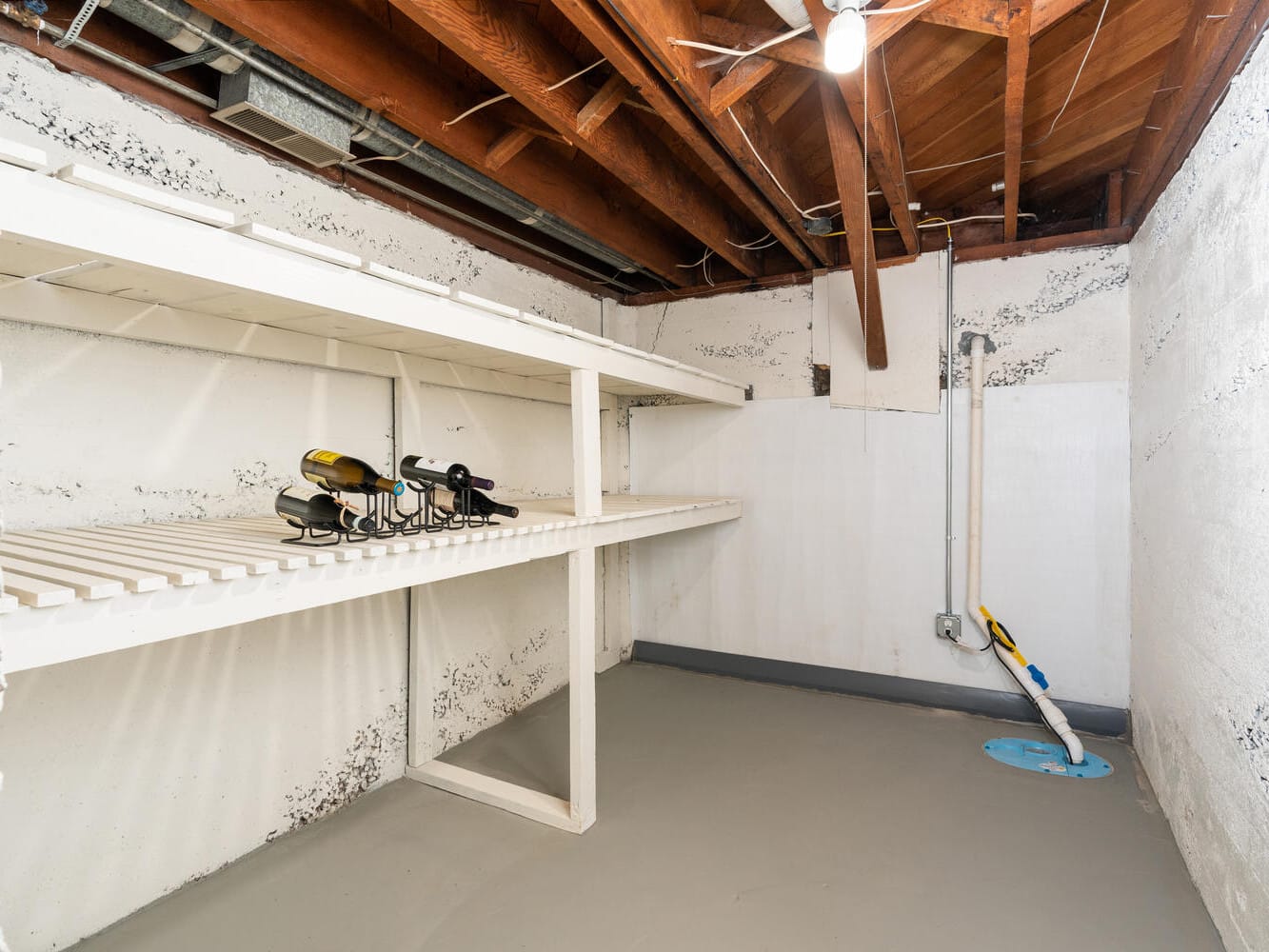 Basement storage room with white shelves on the left, housing some tools. The room has an unfinished wooden ceiling, concrete walls with some visible cracks, and a gray floor with a small blue sump pump in the corner.