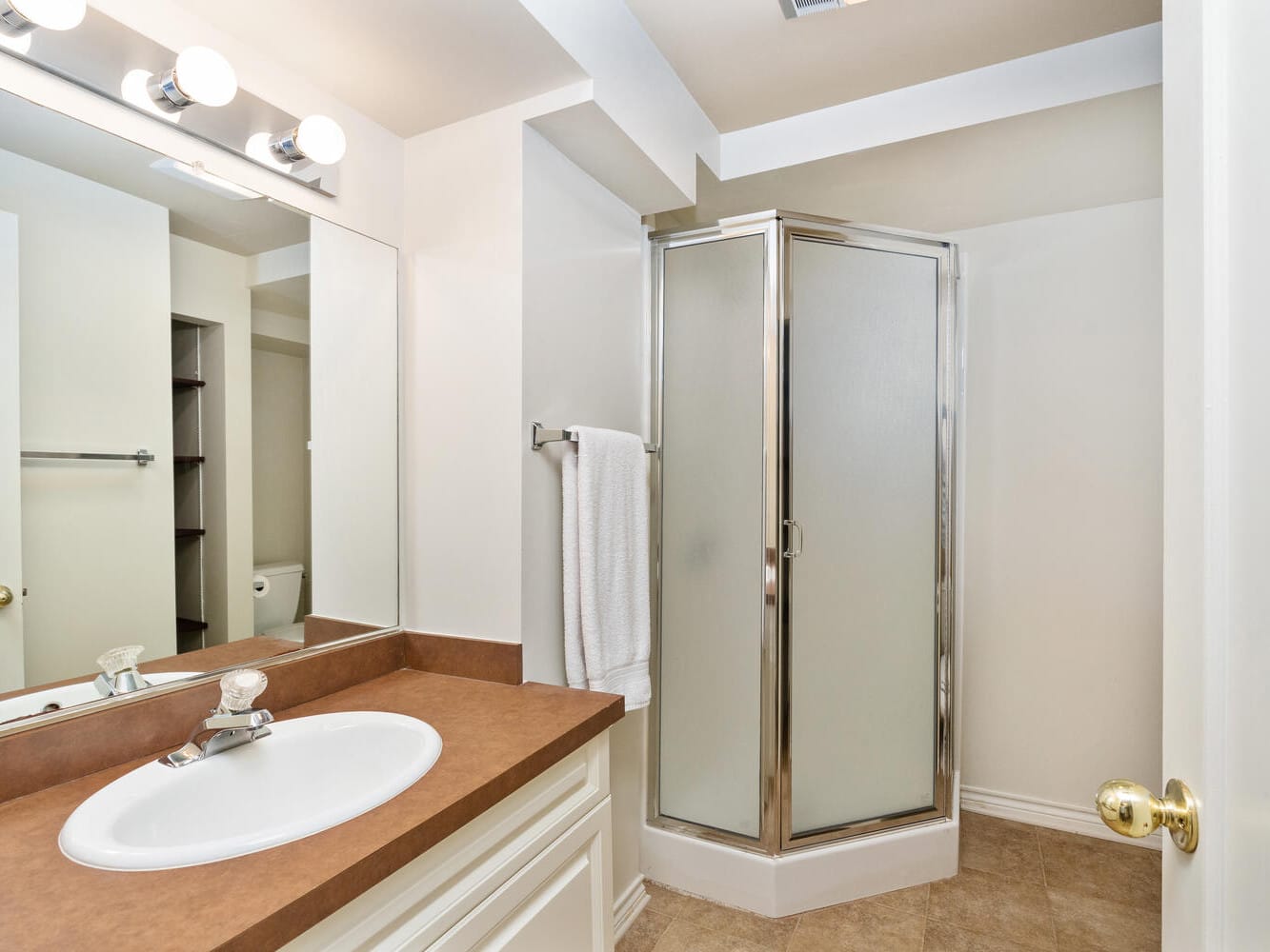 A bathroom with a wood countertop, white sink, and a mirror above. A towel hangs on the metal bar beside a corner shower with frosted glass doors. The floor is tiled and the lighting is bright with three bulbs above the mirror.