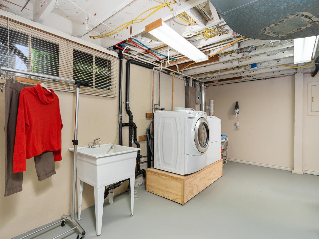 A basement laundry room featuring a washing machine and dryer on a wooden platform, a utility sink, and a rack with clothes. There are exposed pipes and wires on the ceiling and fluorescent lights hanging above.