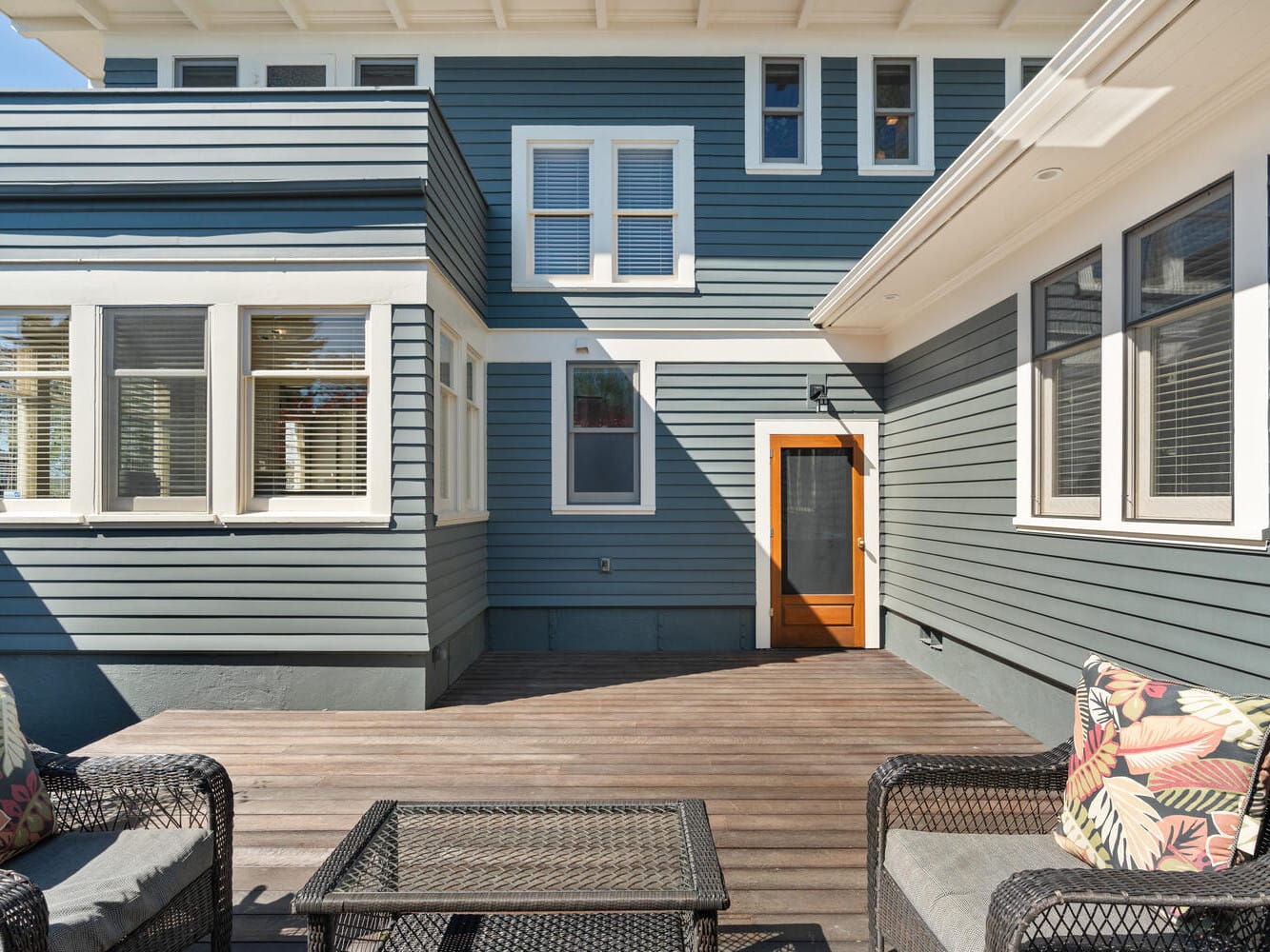 A spacious wooden deck with two wicker chairs and a glass-top table adorned with colorful cushions. The backdrop features a large, blue-gray house with white trim and multiple windows. A wooden door leads indoors.