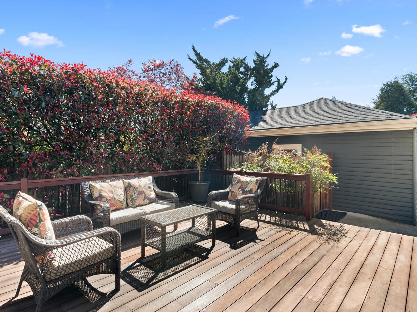 A sunny wooden deck with wicker furniture, including a sofa, two chairs, and a glass-top coffee table. The deck is bordered by lush red and green bushes, and a gray wooden house is visible in the background. Clear blue sky above.