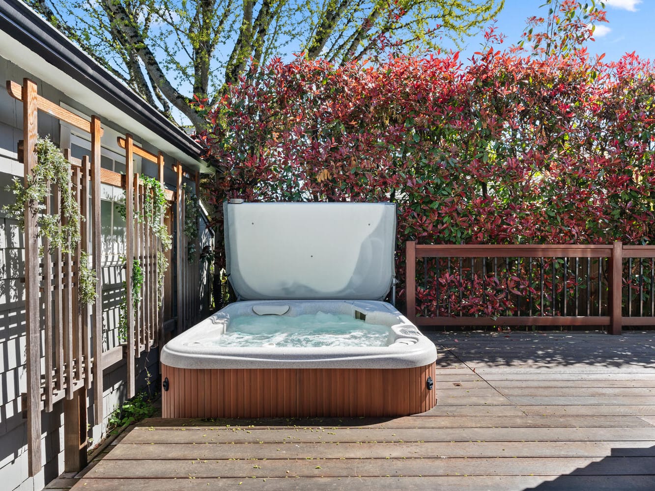 A hot tub sits on a wooden deck, surrounded by a privacy screen and lush red and green foliage. The sky is clear and blue.