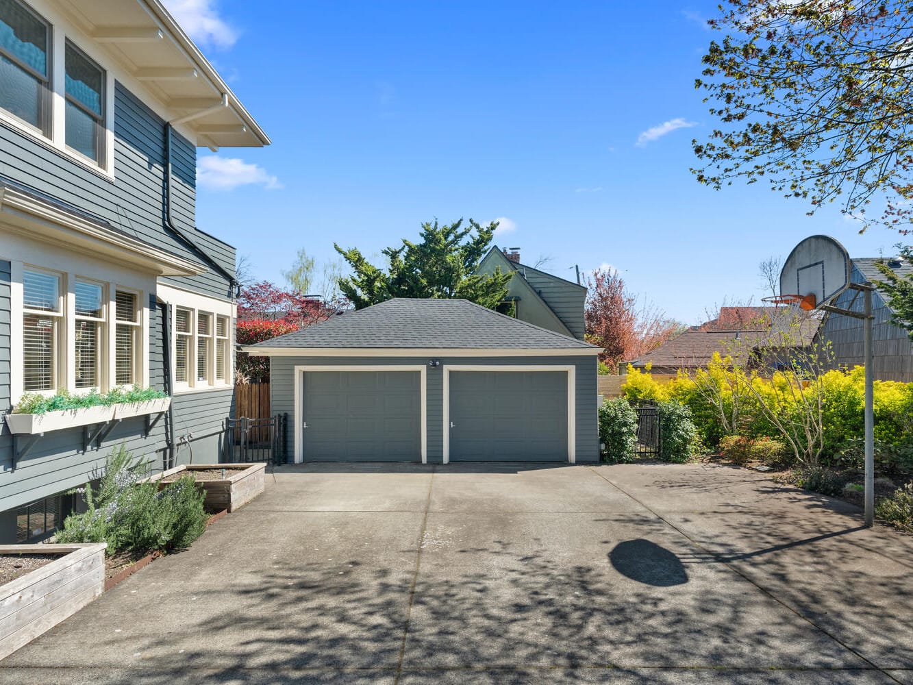 A two-story gray house with white trim and a two-car garage. The front yard features planters and shrubs, with a paved driveway leading to the garage. Trees and a clear blue sky are in the background.