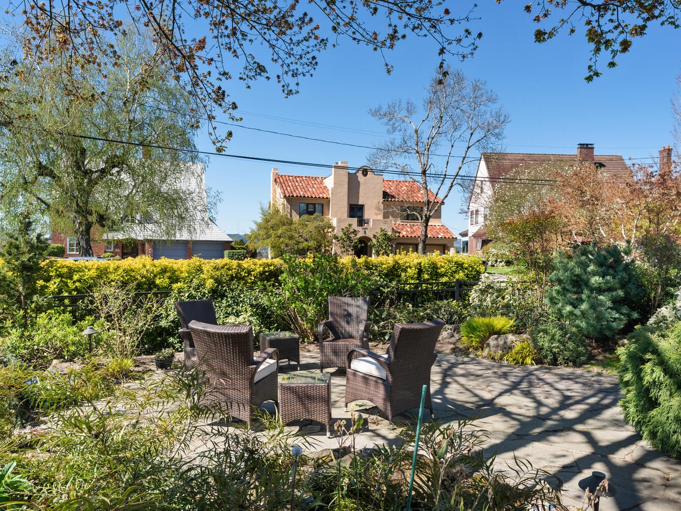 A serene backyard patio features a set of brown wicker chairs and a table surrounded by lush greenery. In the background are two residential houses with red-tiled roofs beneath a clear blue sky. Trees frame the scene.