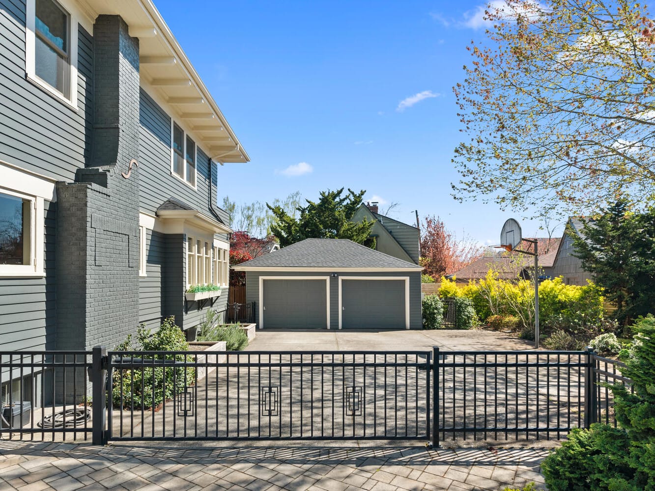 A two-story gray house with white trim features a fenced front yard. A basketball hoop stands beside a paved driveway leading to a detached two-car garage. The yard is landscaped with trees and shrubs, under a clear blue sky.