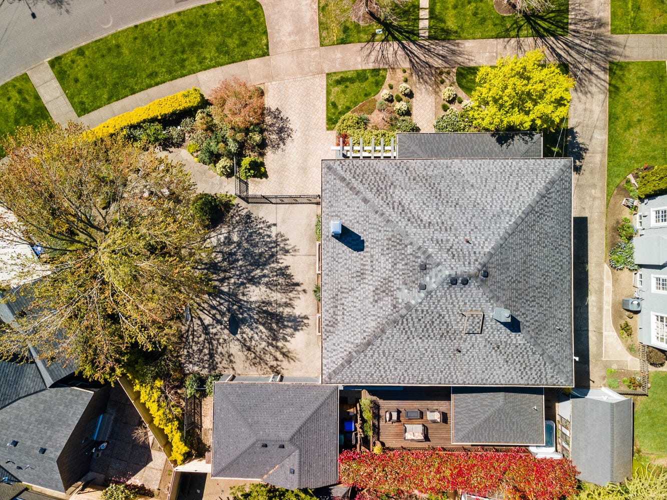 Aerial view of suburban neighborhood showing a house with a gray roof, surrounded by trees and greenery. Adjacent houses and roads are visible, with neatly arranged yards and landscaped areas.