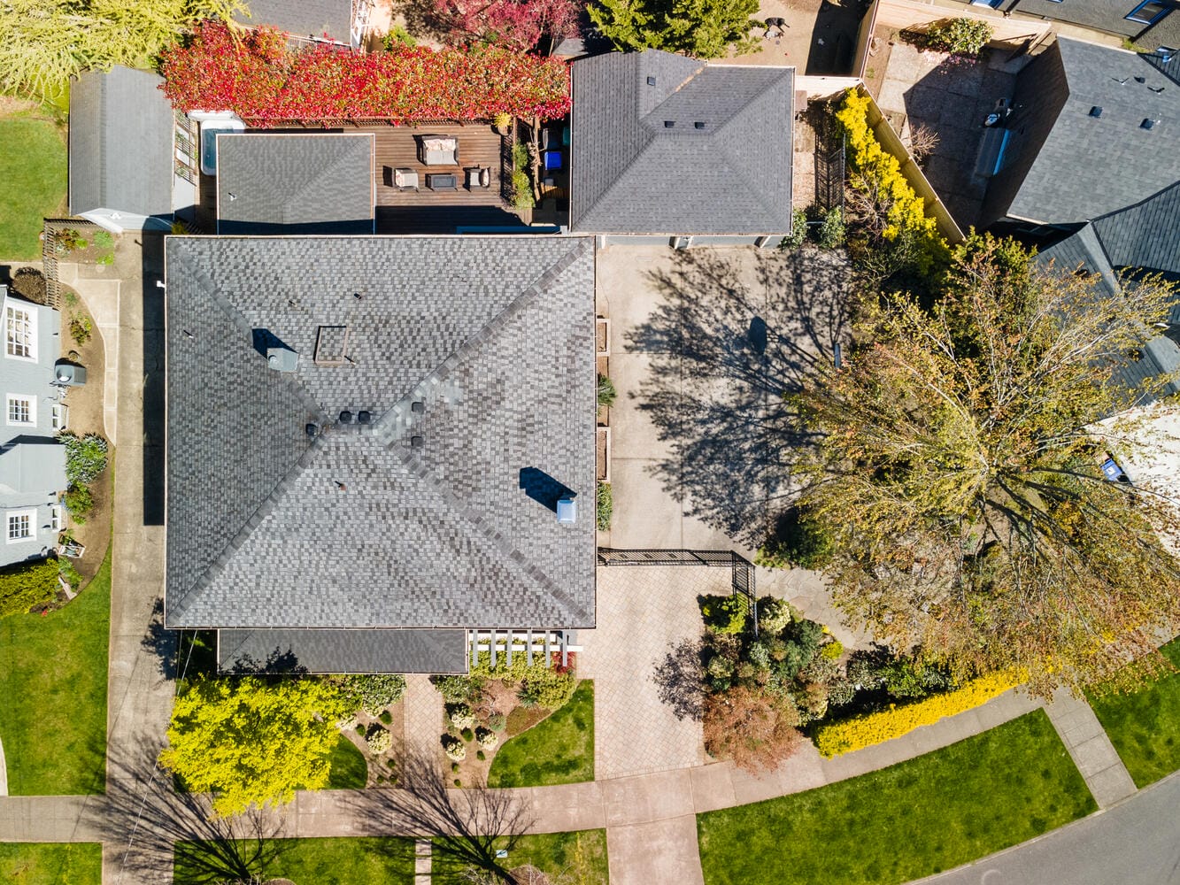 Aerial view of a residential area showcasing a house with a large, dark gray roof and surrounding greenery. A driveway and a small garden with various trees and shrubs are visible. A nearby street curves at the bottom of the image.