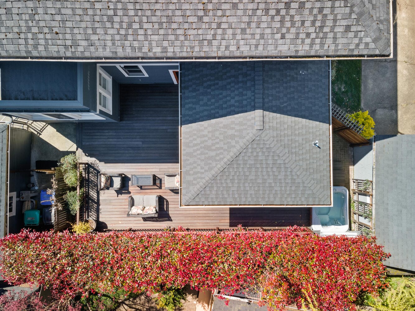 Aerial view of a house with a deck featuring outdoor furniture and a hot tub. The deck is bordered by a vibrant tree with red leaves, situated between two rooftops. Surfaces vary between shingles and wooden planks.