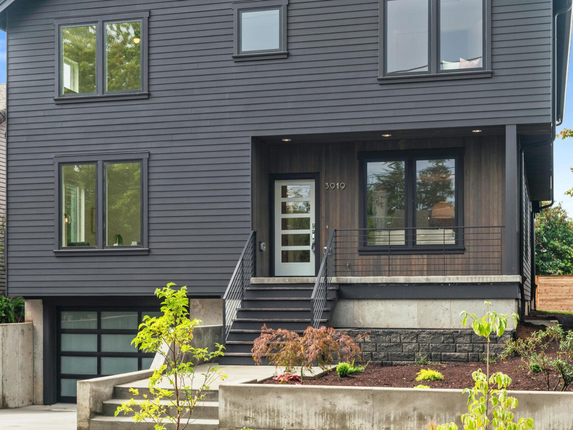 A modern two-story house with dark gray siding and large windows. The entrance features a glass front door, steps leading up, and a small porch with a railing. Theres a garage below and landscaped greenery in the front yard.