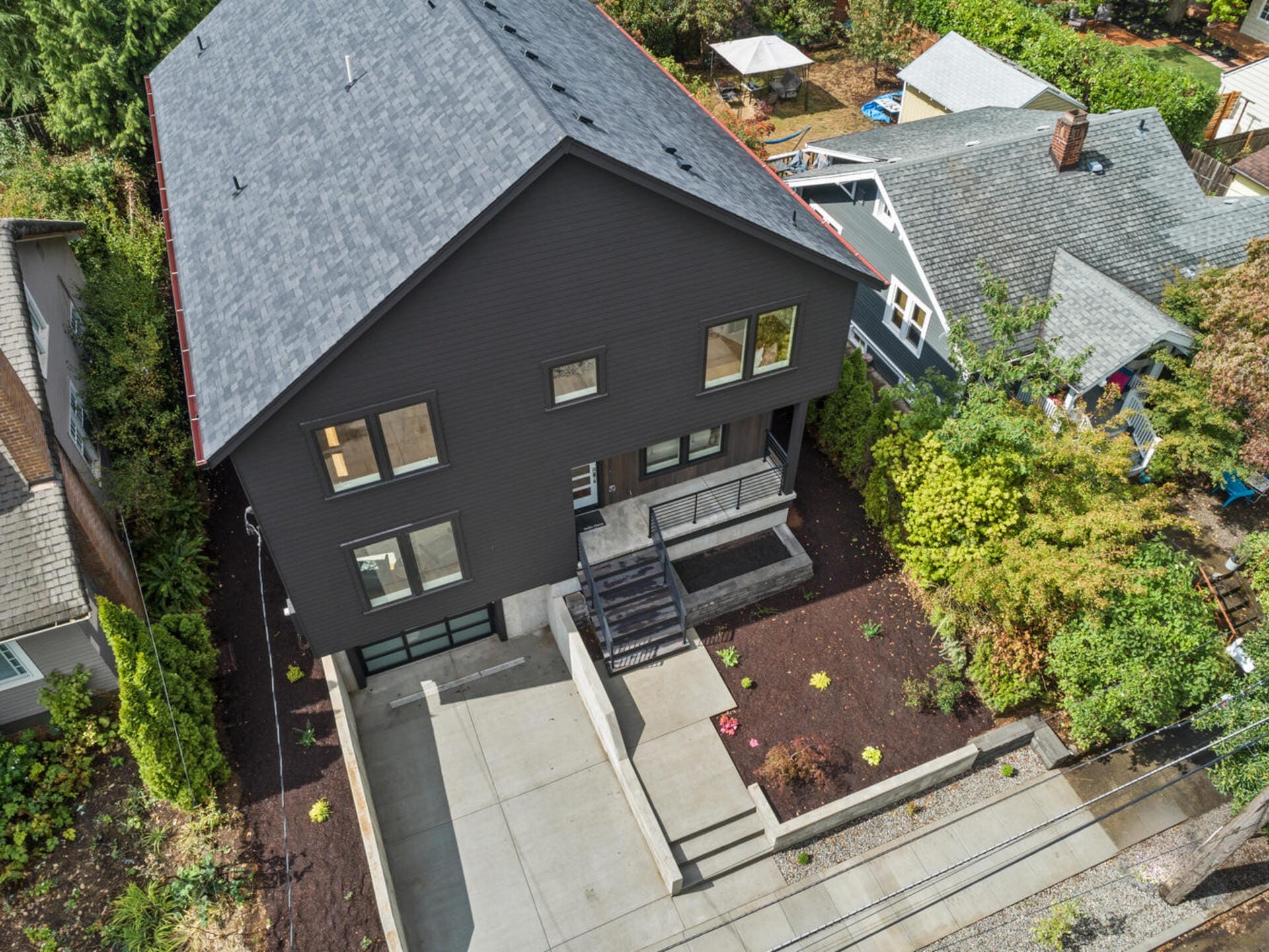 Aerial view of a modern two-story house with dark siding and a gray roof. It features large windows, a spacious concrete driveway, and landscaped garden beds. The house is surrounded by greenery and neighboring homes.