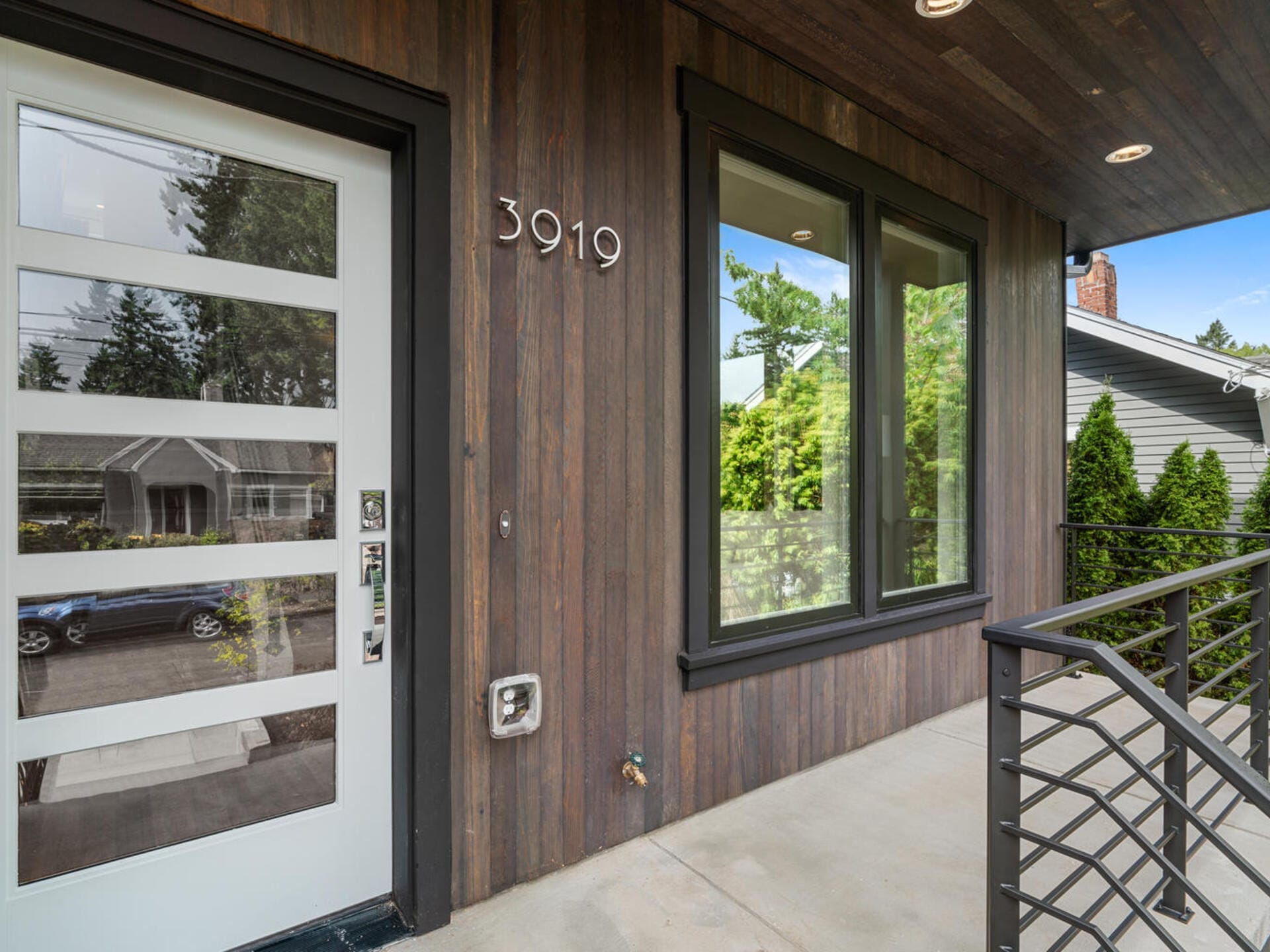 Modern front porch with dark wood siding, a white door featuring frosted glass panels, and large windows reflecting greenery. The house number 3919 is visible beside the door. A metal railing lines the edge of the porch.