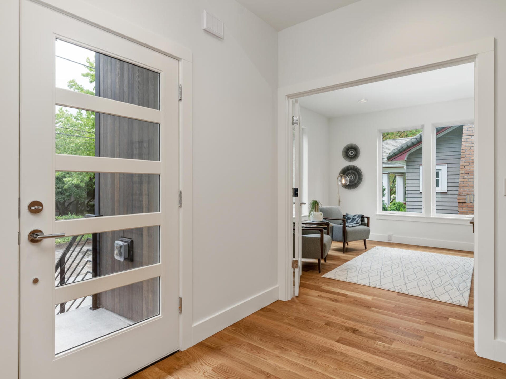 A modern entryway with a glass-paneled door leading to a room with wooden flooring. The adjacent room has a large window, white walls, decorative plates on one wall, and a gray armchair. A small table sits on a patterned rug.