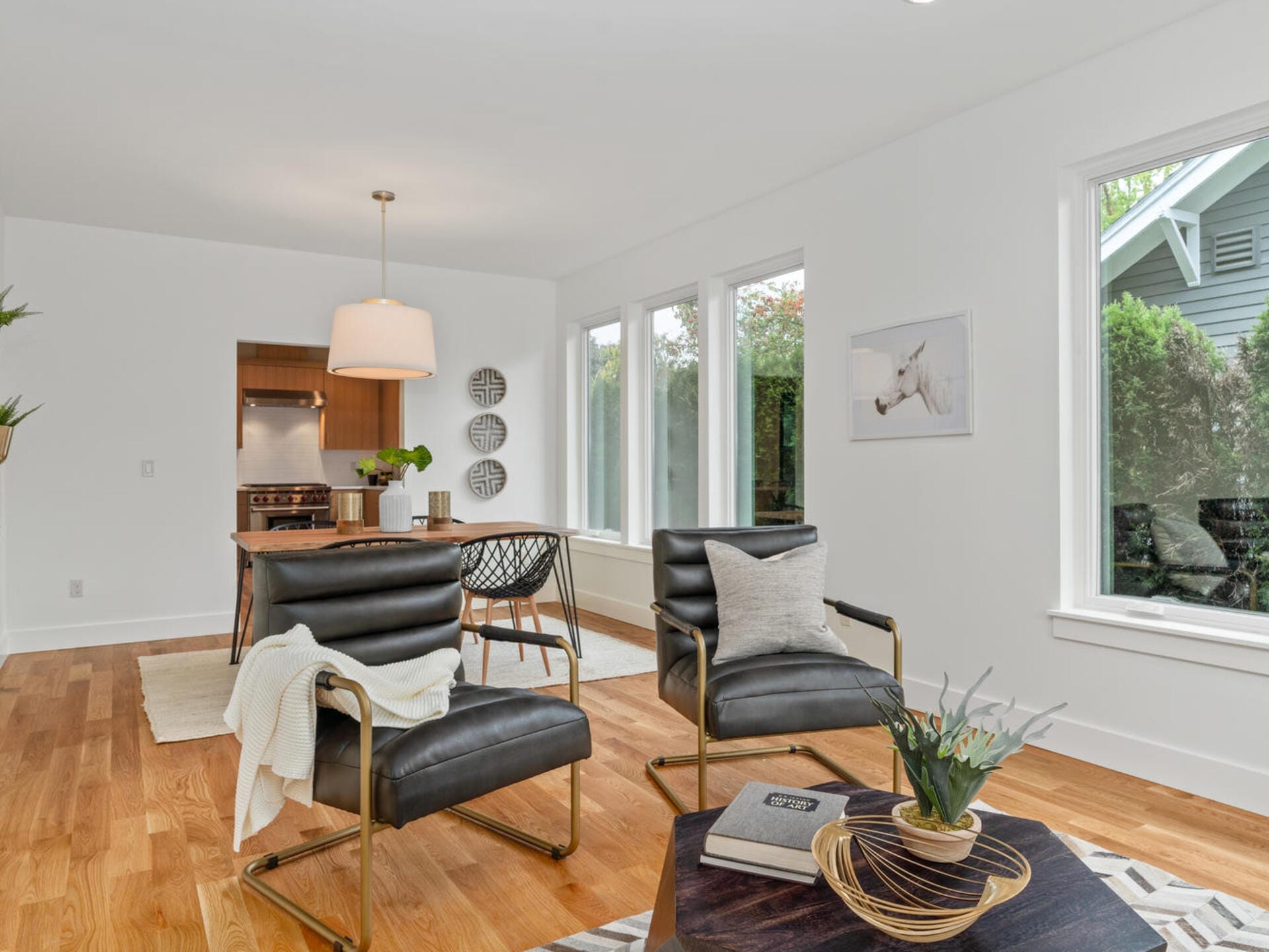 A modern living room with two black leather chairs, a wooden coffee table, and a dining area in the background. The room features hardwood floors, large windows, a pendant light, and minimal decor, including a framed artwork on the wall.