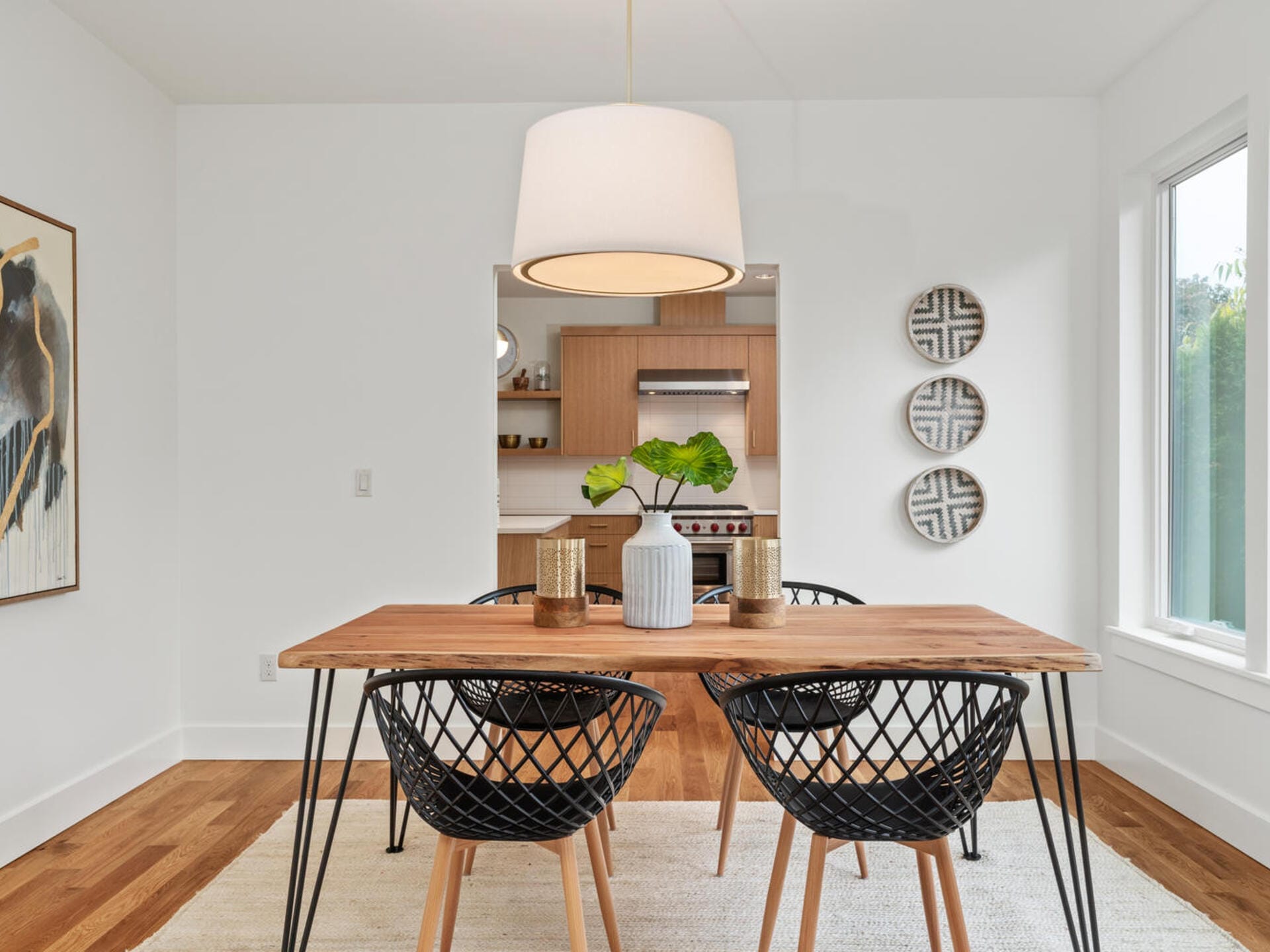 A modern dining room with a wooden table and four black lattice chairs. A large white pendant light hangs above. The room features a neutral color palette with wall art and decorative plates, and has a view into a kitchen with wooden cabinets.
