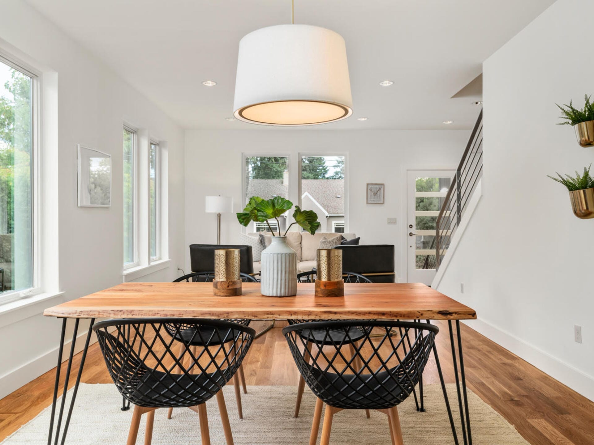 A modern dining area with a wooden table, two black woven chairs, and decorative vases. Large windows bring in natural light, highlighting the wooden floor. A white pendant lamp hangs above the table. Green plants add a touch of nature.