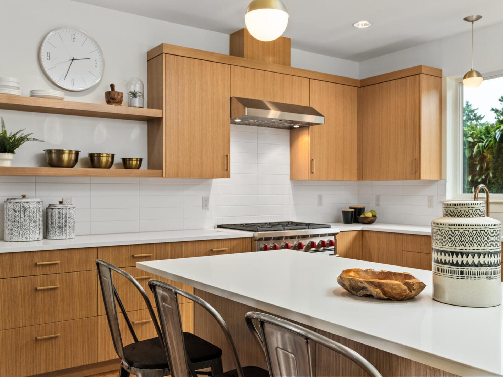 Modern kitchen with light wood cabinets, white countertops, and a white tile backsplash. A clock hangs above open shelving with decor items. Stainless steel stove with range hood, and a large island with metal chairs and decorative bowls.