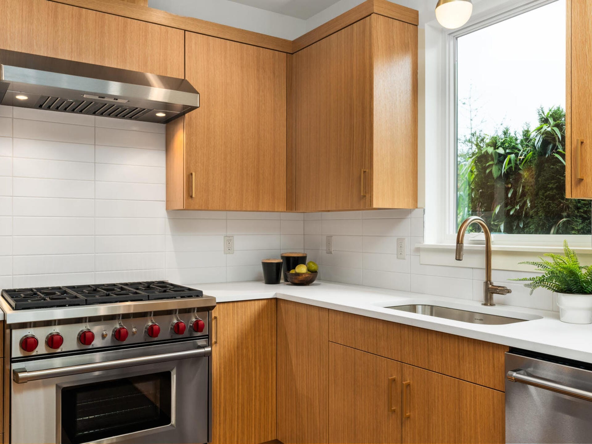 A modern kitchen with wooden cabinets, a stainless steel gas stove with red knobs, a sink with a faucet, a window, and a white tiled backsplash. A potted plant and a tray with fruit are on the white countertop.