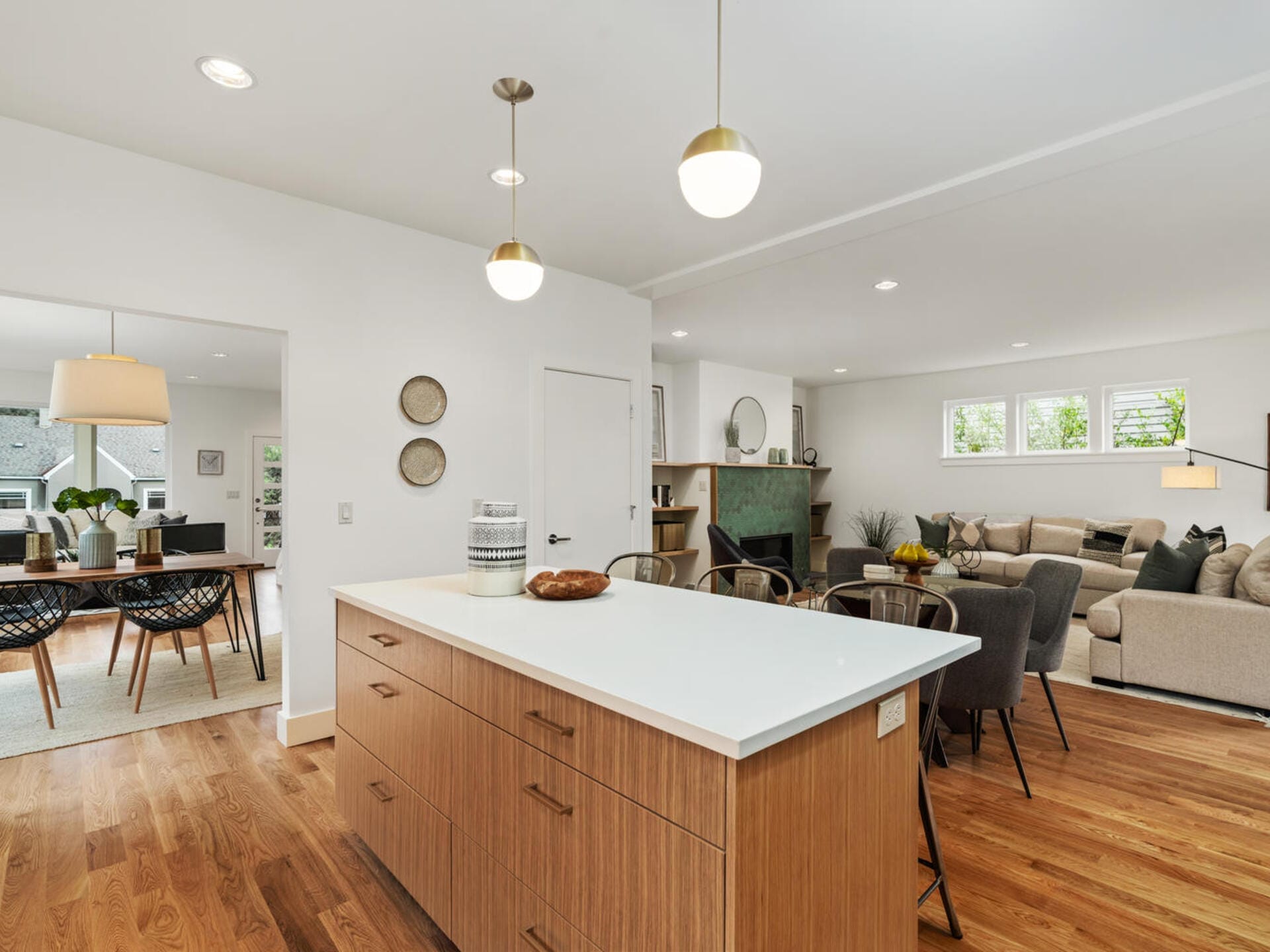 A modern open-concept kitchen and living room. The kitchen features a wooden island with a white countertop and pendant lights. In the background, there is a dining area with a table and chairs, and a cozy living space with a sofa and armchairs.