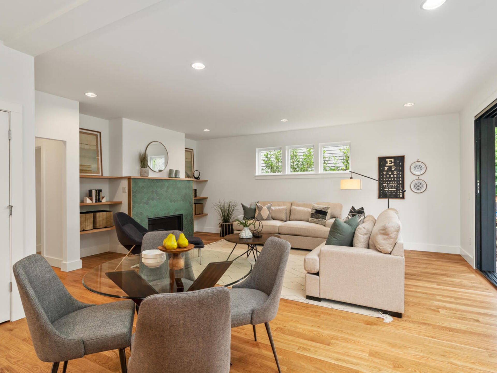 Spacious living room with modern decor, featuring a green-tiled fireplace, beige sofas, and a round glass dining table with gray chairs. The room has wooden flooring, wall art, and large windows allowing natural light. A small desk area is visible.