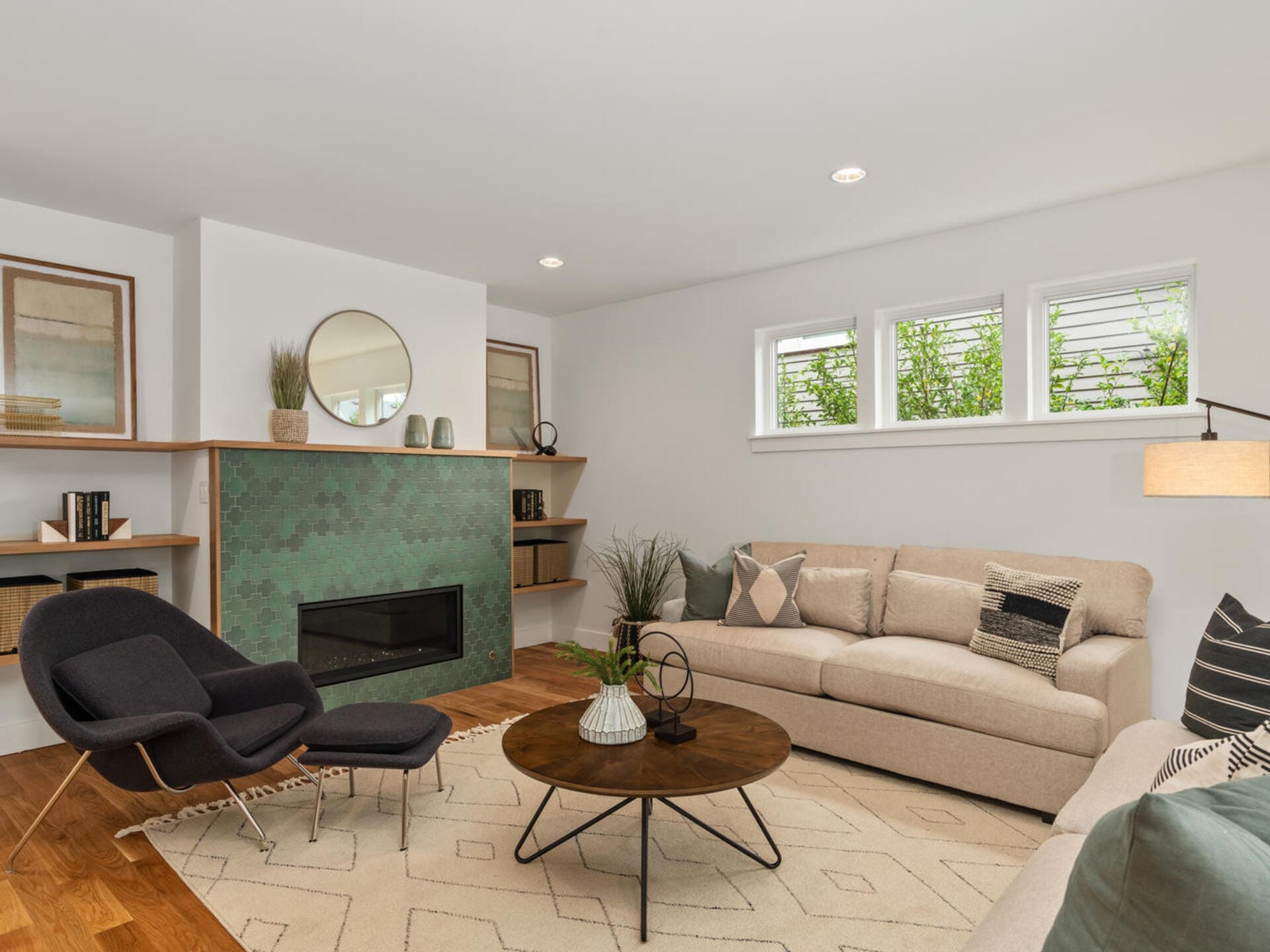A cozy living room with a beige sectional sofa, black lounge chair, and round wooden coffee table. The green-tiled fireplace features a round mirror above it. Shelves on each side hold books and decor. Three small windows add natural light.