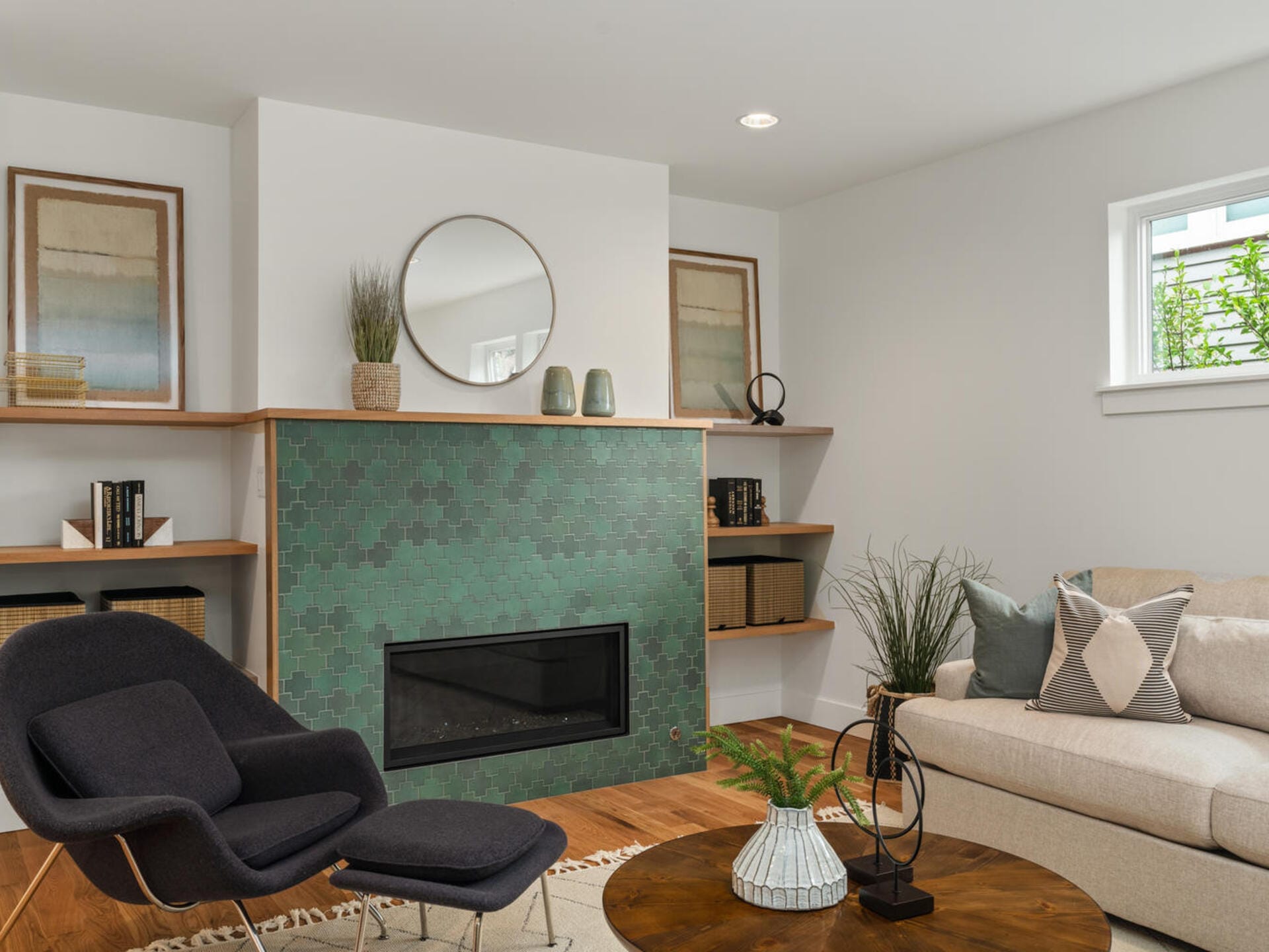 A modern living room featuring a green tiled fireplace with a round mirror above it. Shelves on both sides hold books and decor. A black chair and beige sofa surround a wooden coffee table with plants and ornaments. Natural light filters through a window.