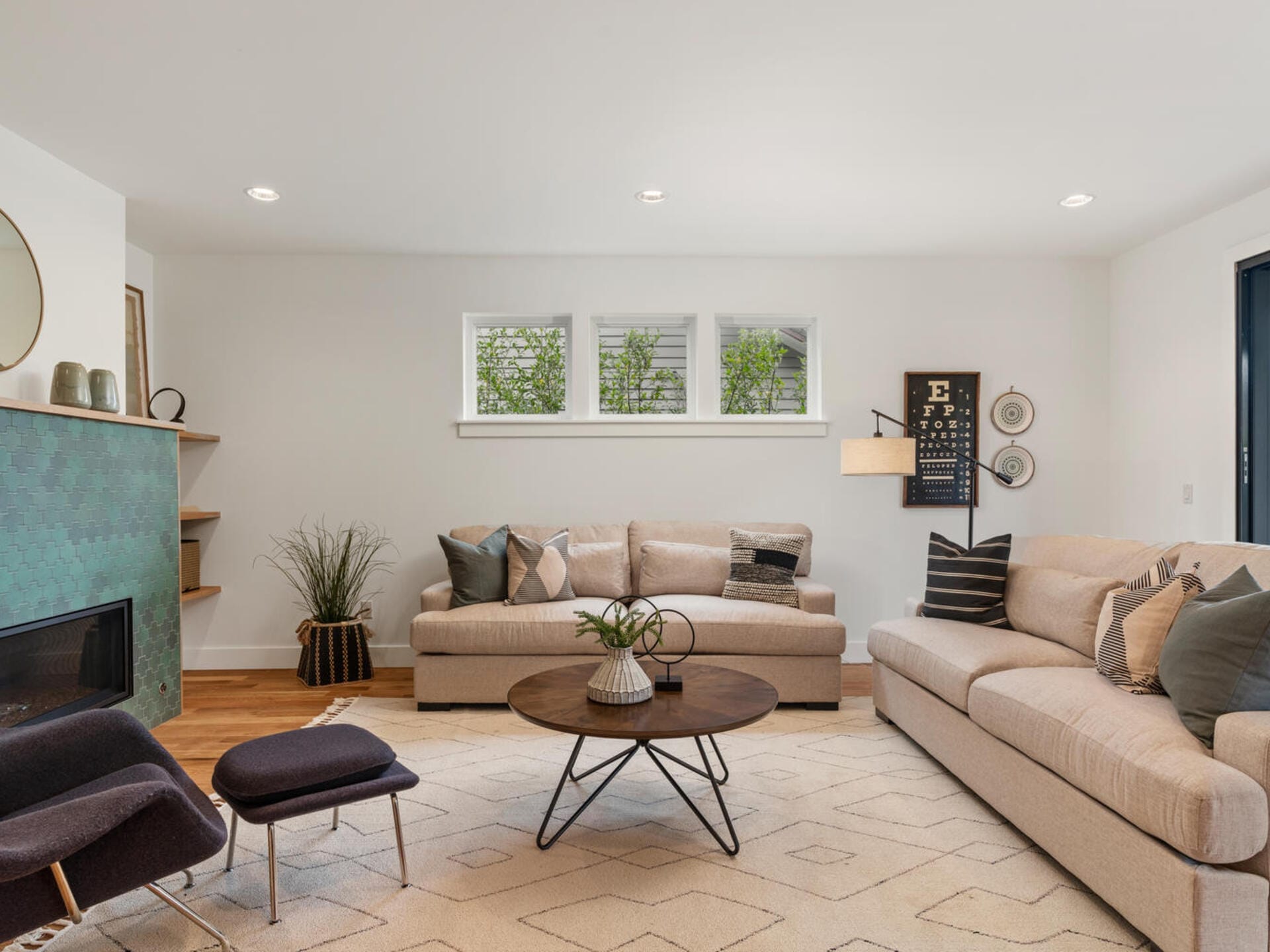 A modern living room with two beige sofas, a round wooden coffee table, and an accent chair. The room features a green-tiled fireplace, a geometric rug, and wall decor. Three small windows provide natural light. Neutral and green tones dominate the space.