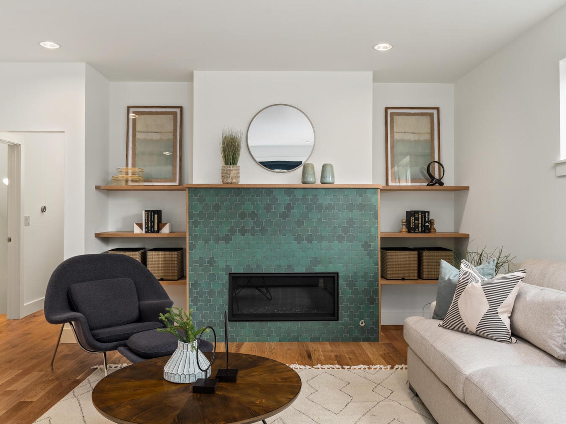 A modern living room with a green hexagonal-tiled fireplace, a circular mirror above, and wooden shelves with decorative items on either side. A gray chair, a beige sofa with patterned cushions, and a round wooden coffee table complete the space.