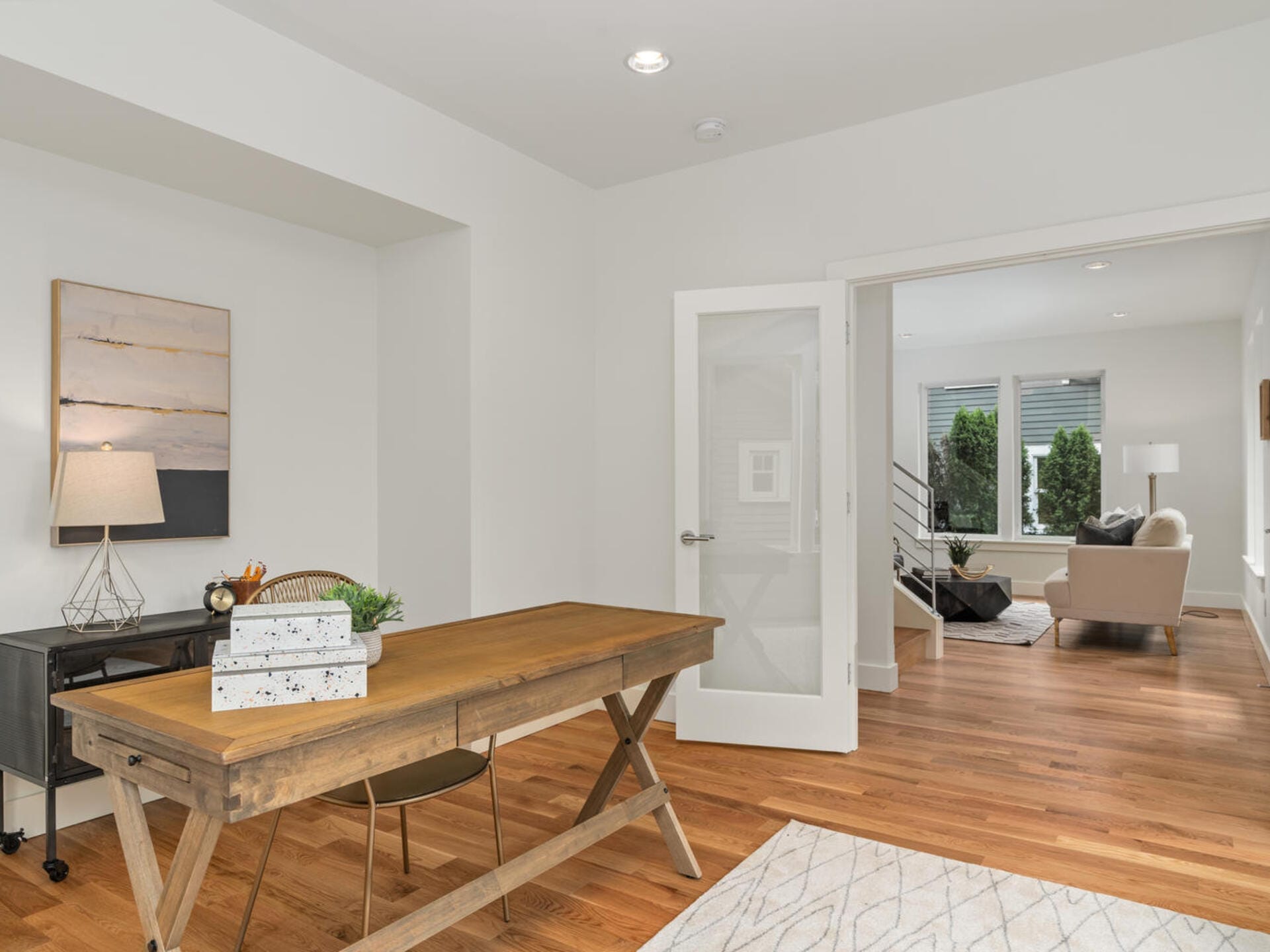A bright, modern home office with a wooden desk and chair on a wooden floor. A lamp and book sit on the desk. A large painting hangs on the wall. Through an open door, a living room with a sofa and large windows is visible.