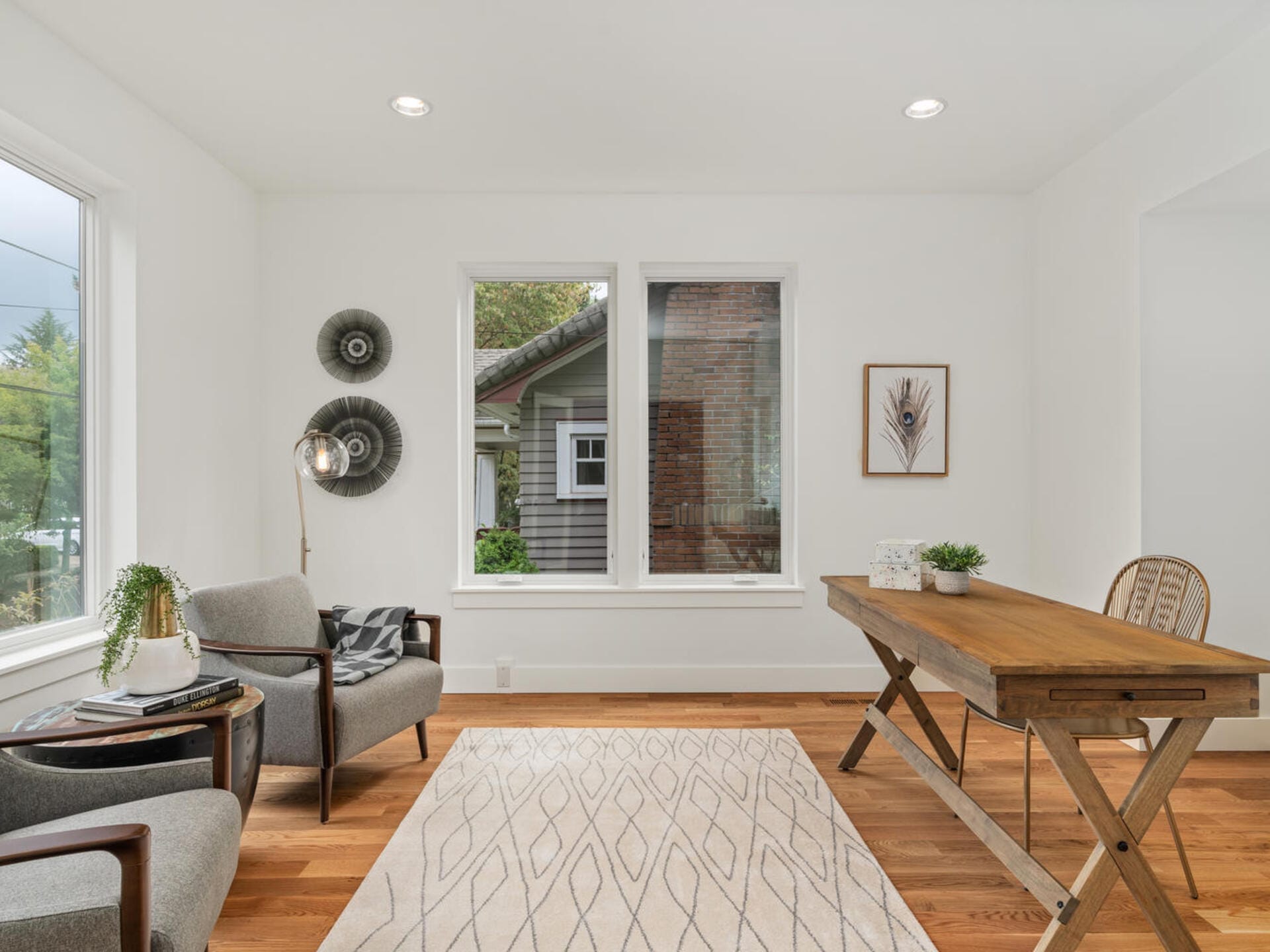 A modern room with a wooden desk and a wicker chair on the right, two gray armchairs on the left, and a geometric-patterned rug on the wooden floor. Large windows let in natural light, and decorative wall items enhance the minimalist decor.
