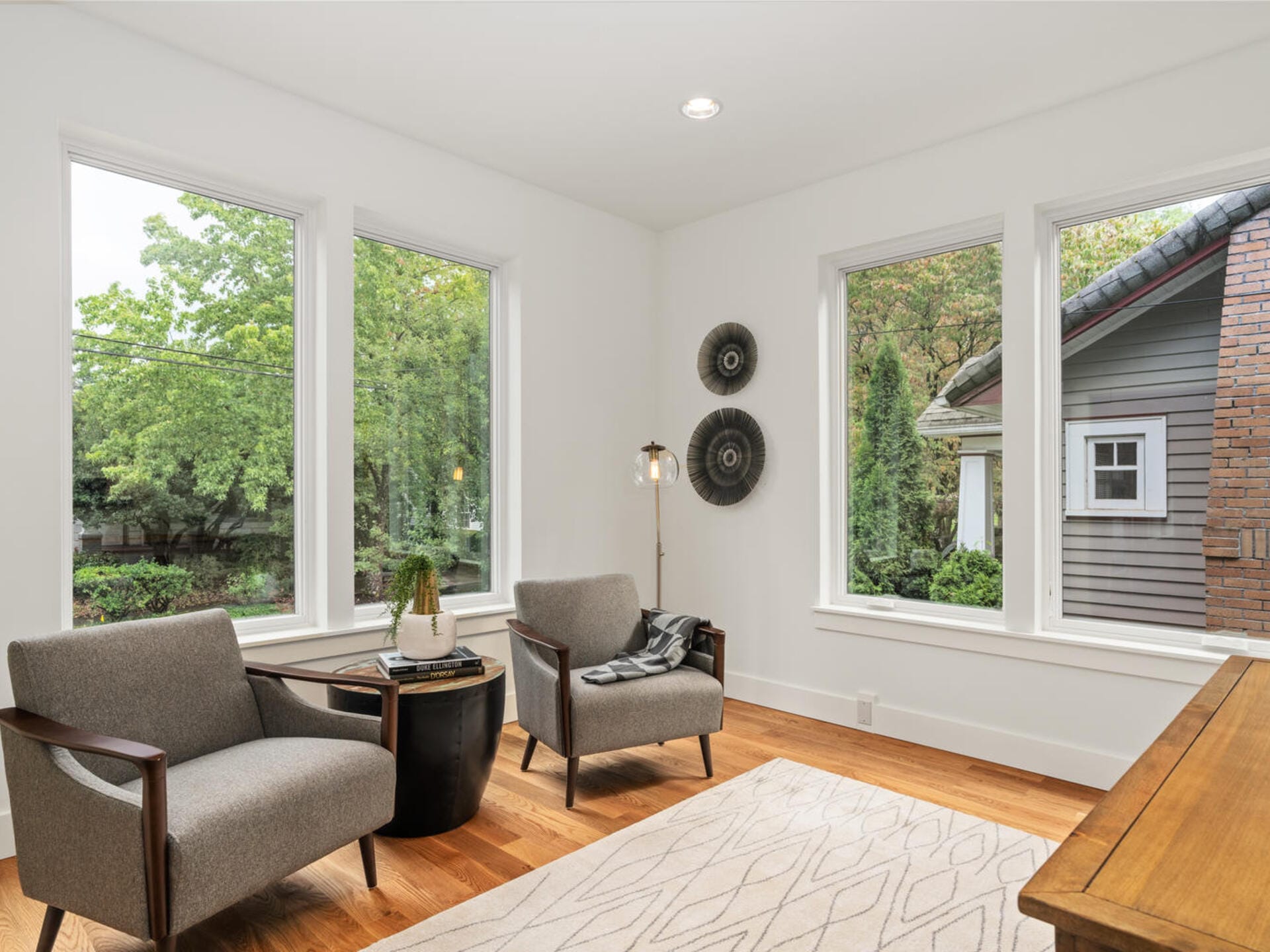 A cozy living room with two gray armchairs and a small round table on a hardwood floor. Large windows offer a view of greenery outside, and a patterned rug lies on the floor. Two circular wall decorations add a touch of style to the room.