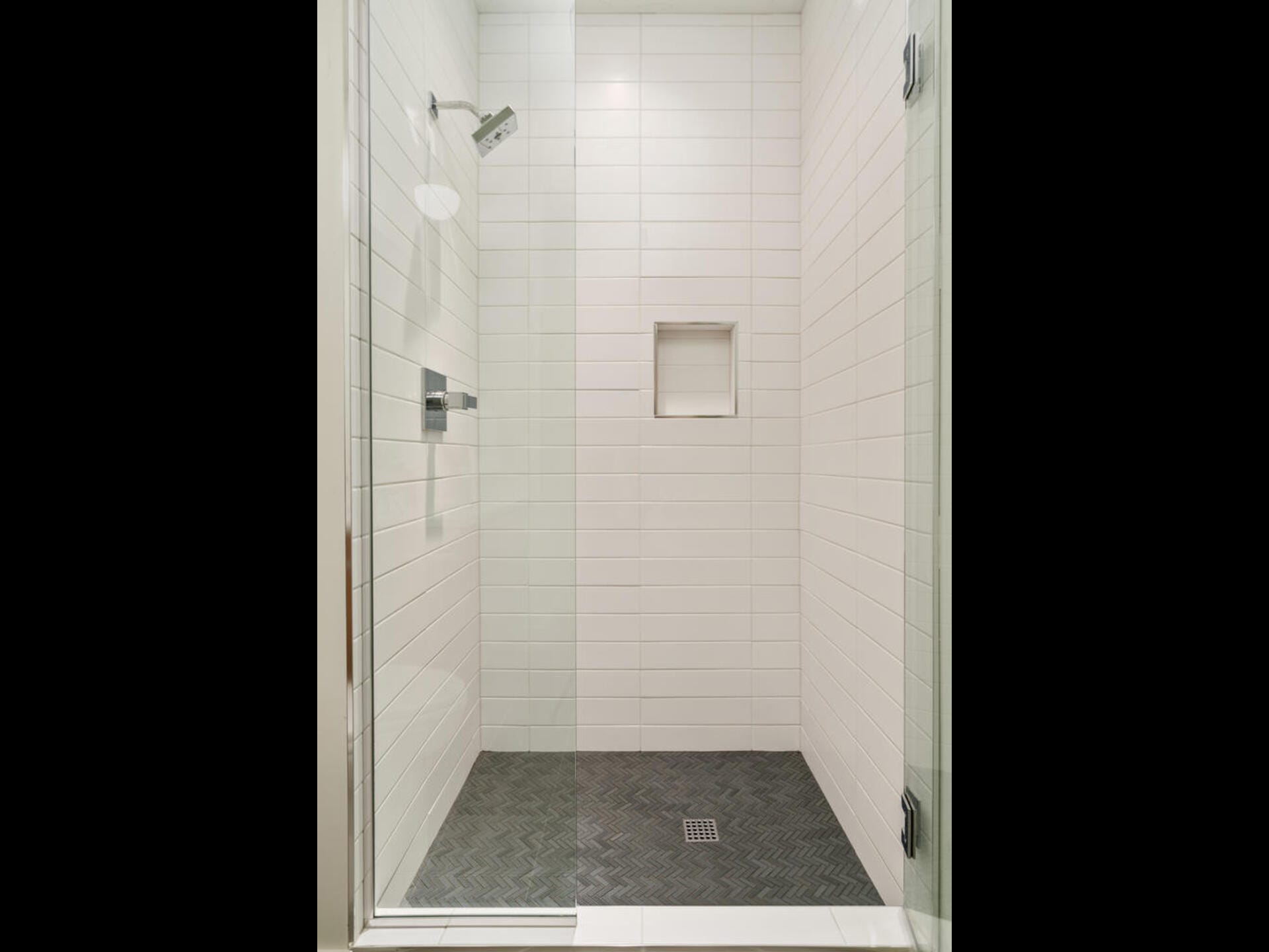 A spacious, modern shower with white subway tiles, a glass door, a showerhead, a built-in niche, and a herringbone-patterned dark floor.