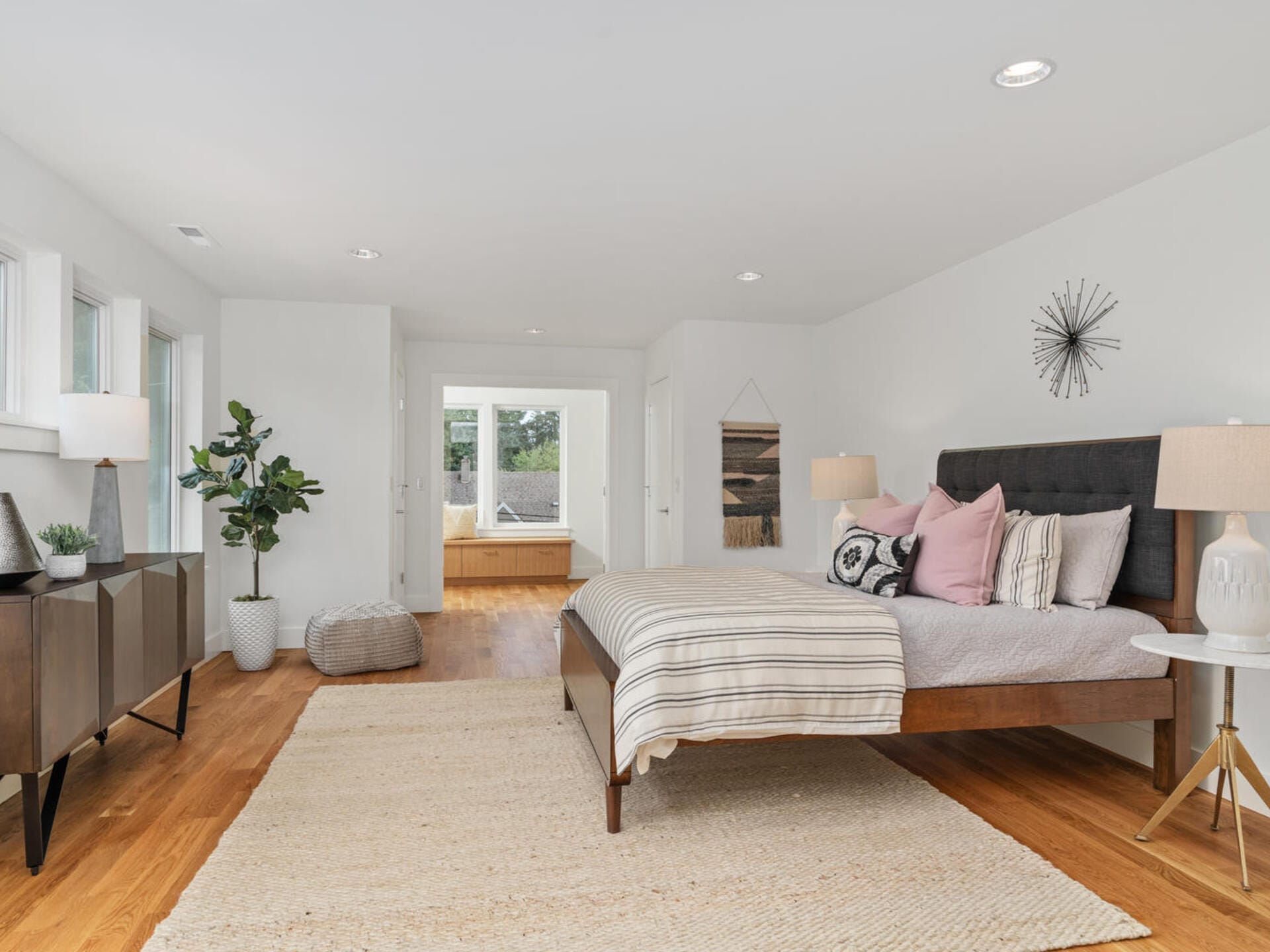 A modern, spacious bedroom with a large bed adorned with pillows and a striped blanket. Wooden flooring is partially covered by a woven rug. The decor features plants, a wall clock, and a sideboard with lamps, creating a cozy atmosphere.