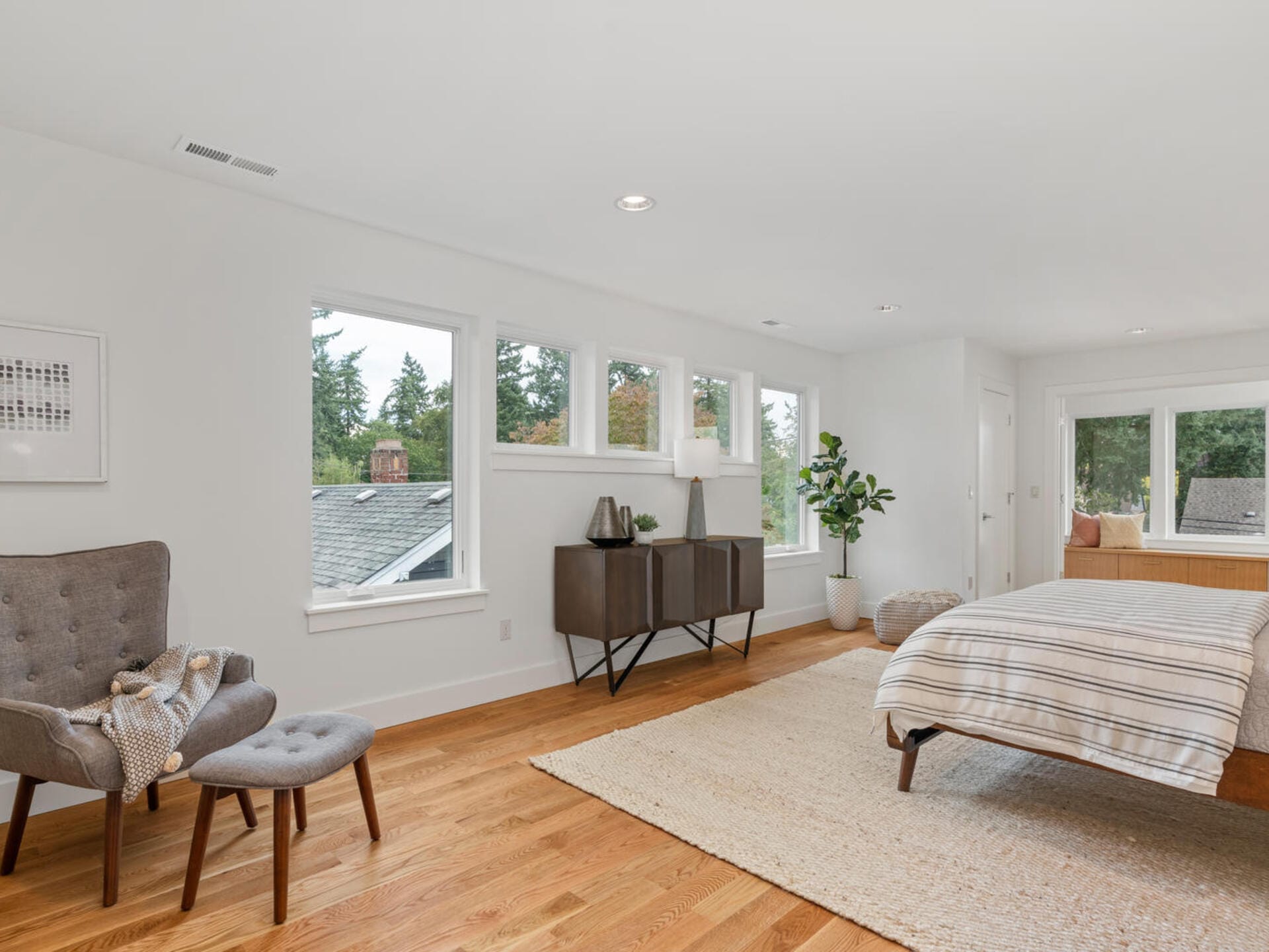 A spacious bedroom with wood flooring, a bed on a light rug, and a cozy gray chair with a blanket. Large windows offer natural light and views of trees. A dark wooden sideboard and a potted plant complete the decor.