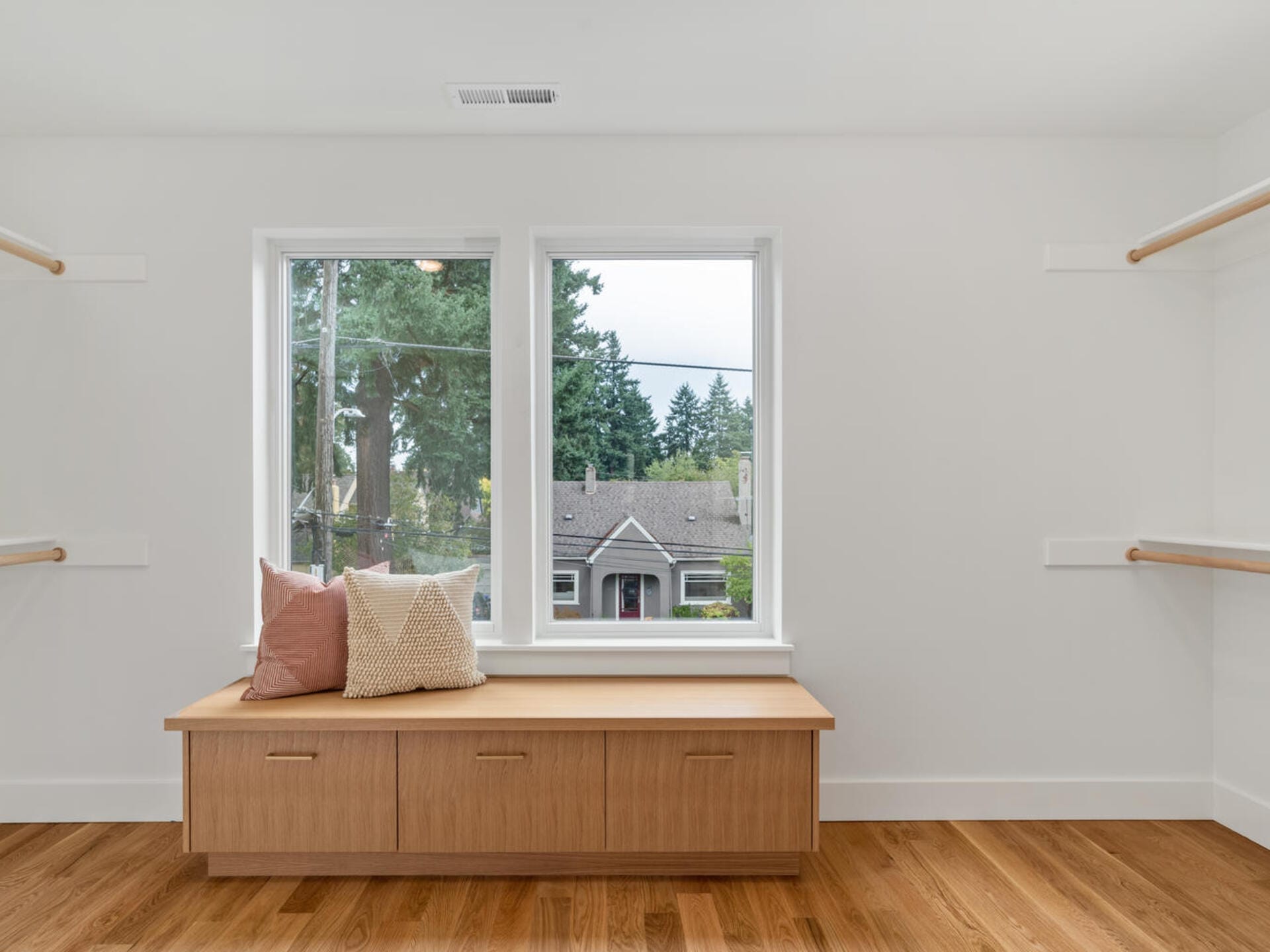 A minimalist room features two large windows with a view of trees and a house. Below the windows, a wooden bench with storage is adorned with two decorative cushions. The room has wooden floors and white walls with light wooden accents.
