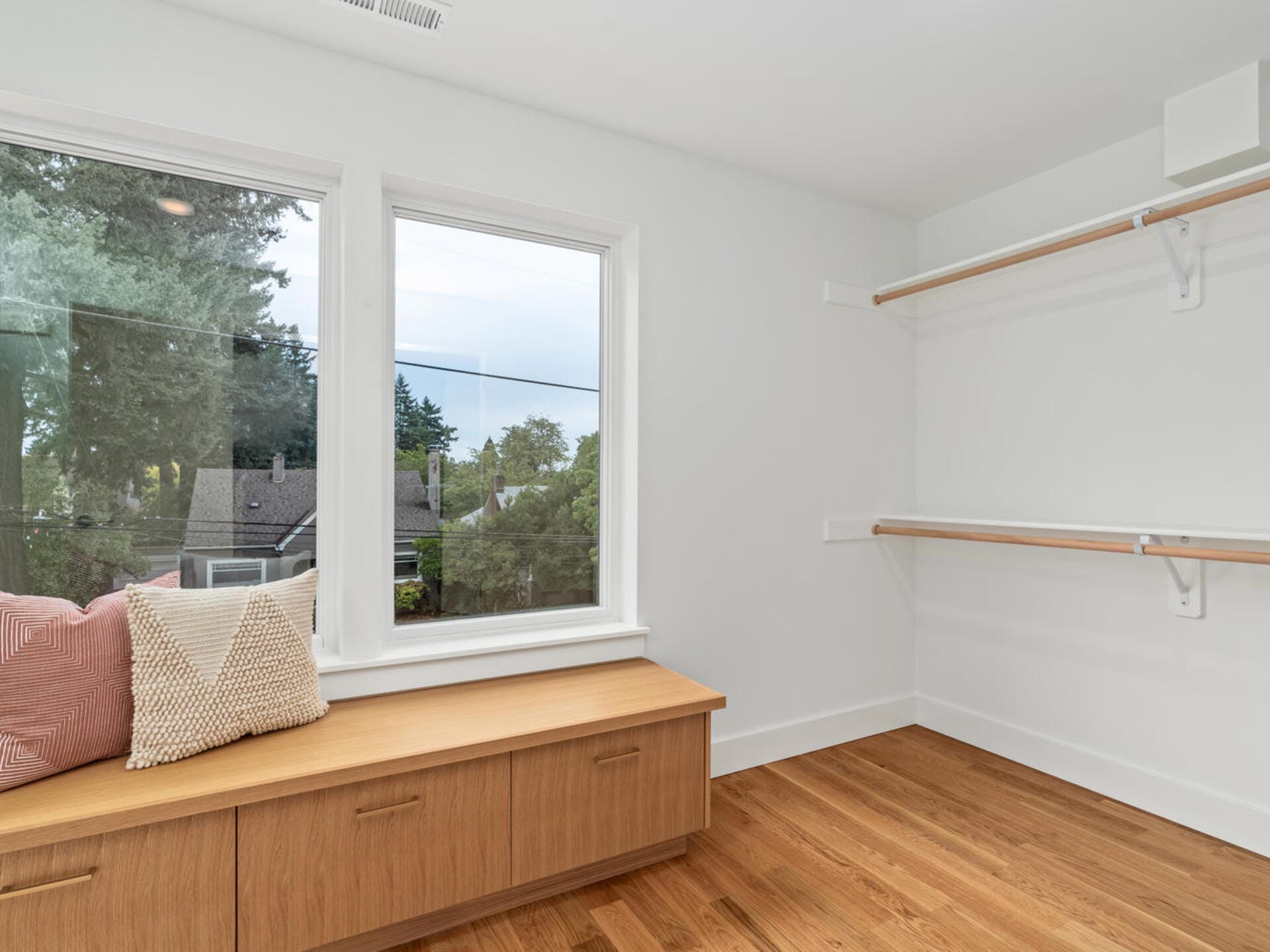 A minimalist room with wooden flooring features a built-in wooden bench with cushions under a large window. The view outside shows trees and rooftops. Two shelves are mounted on the adjacent wall.
