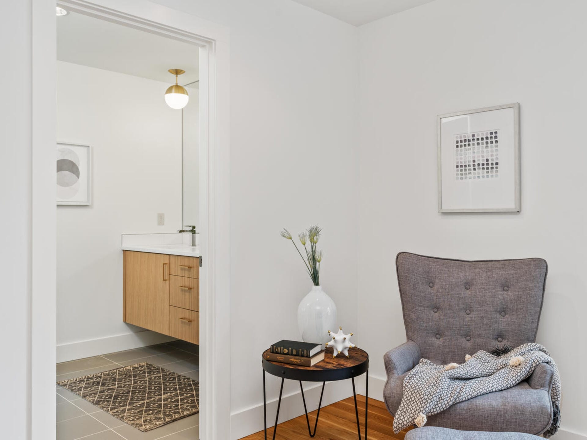 A cozy room with a gray armchair and a small side table featuring a white vase and decorative items. A blanket drapes over the chair. In the background, a bathroom with a wooden cabinet and white walls is visible.