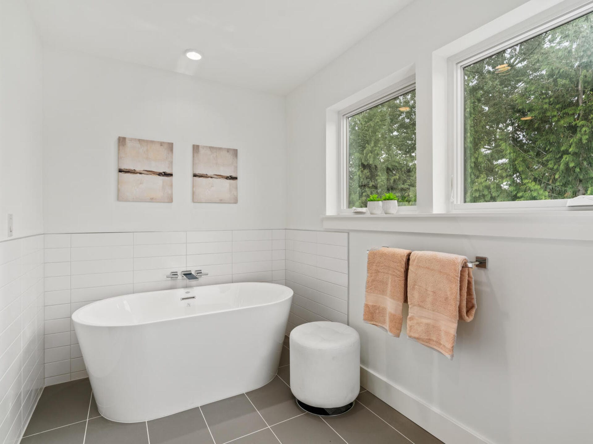 A minimalist bathroom features a white freestanding bathtub adjacent to a small white stool. Two large windows let in natural light. On the wall, two abstract artworks hang above the tub. A towel rack holds two beige towels.