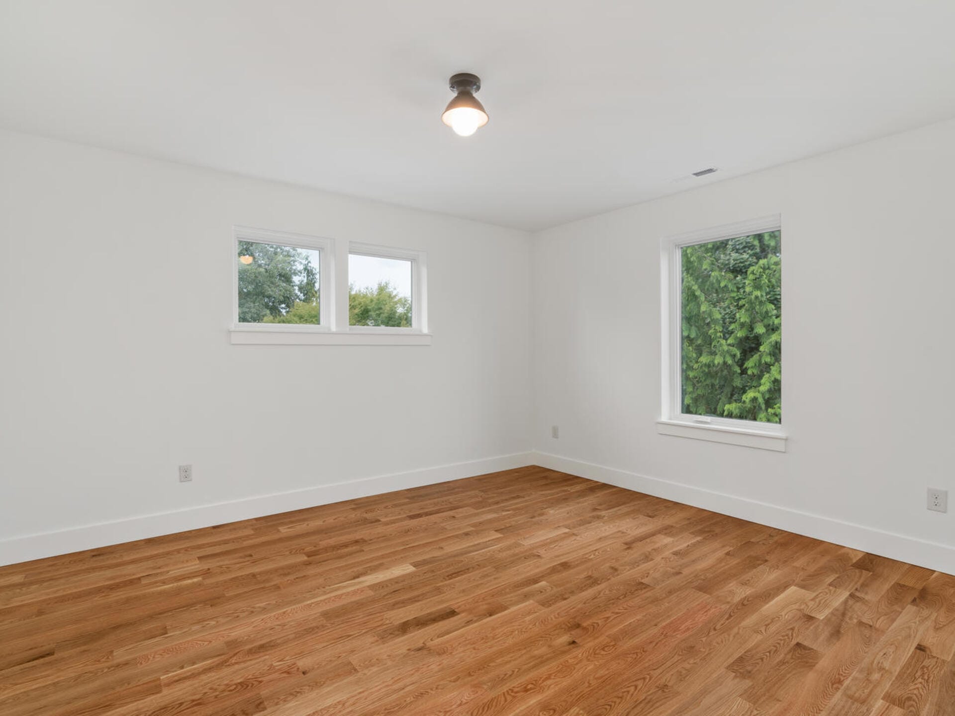A bright, empty room with white walls, wooden flooring, and a single overhead light. Two windows on the left and a larger window on the right bring in natural light, offering views of greenery outside.