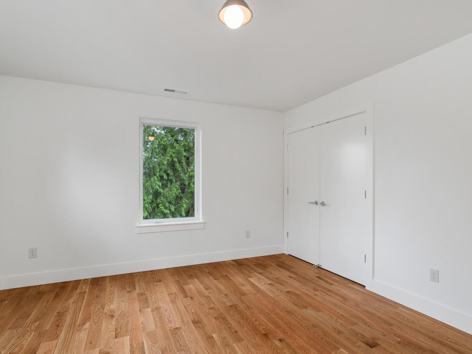 An empty room with white walls and a wooden floor. There is a single window on the left wall with a view of green trees. A closed white double door is on the right wall, and a ceiling light fixture is centered above.