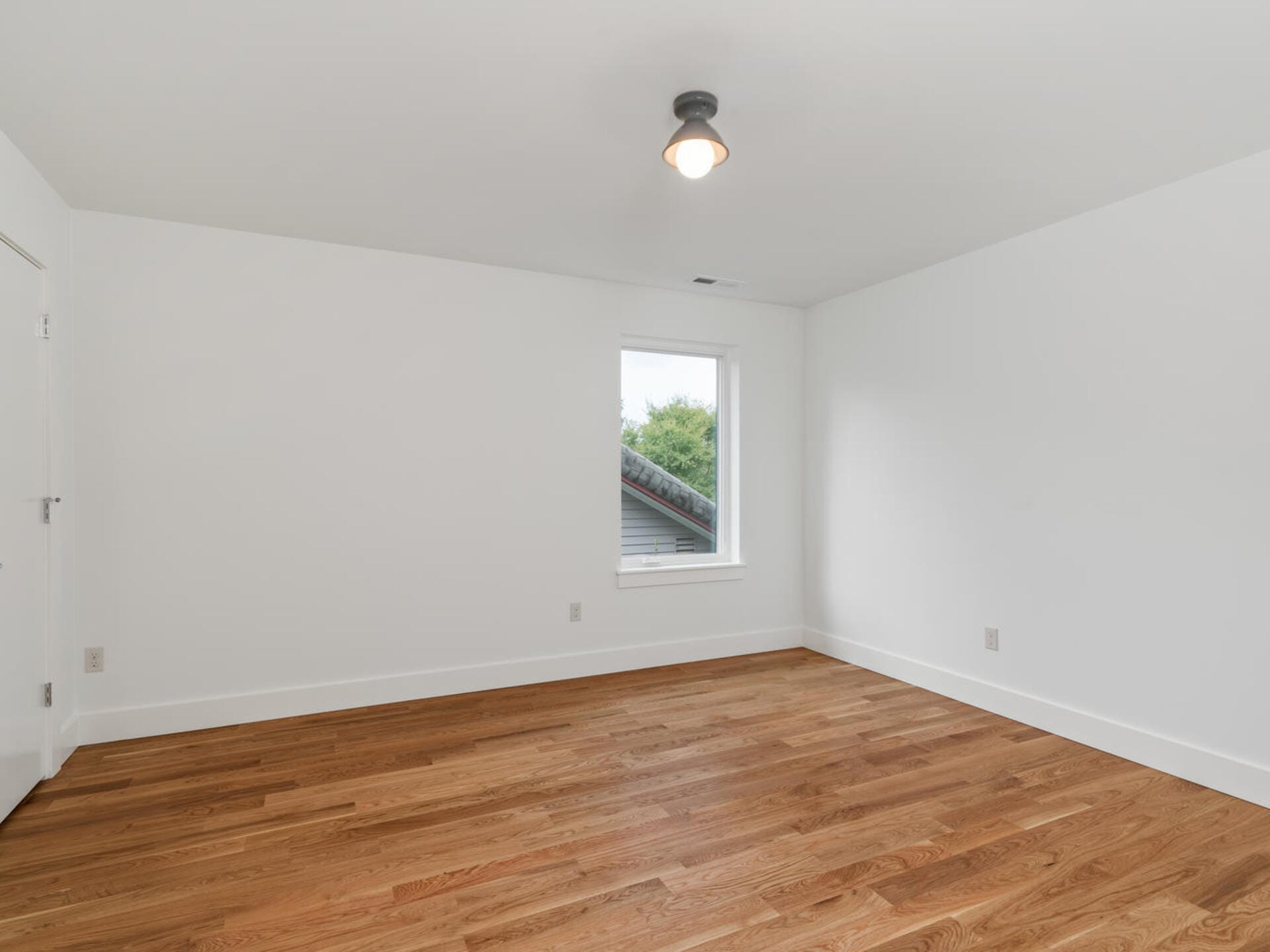 An empty room with white walls, a single window revealing greenery outside, a wooden floor, and a ceiling light fixture.